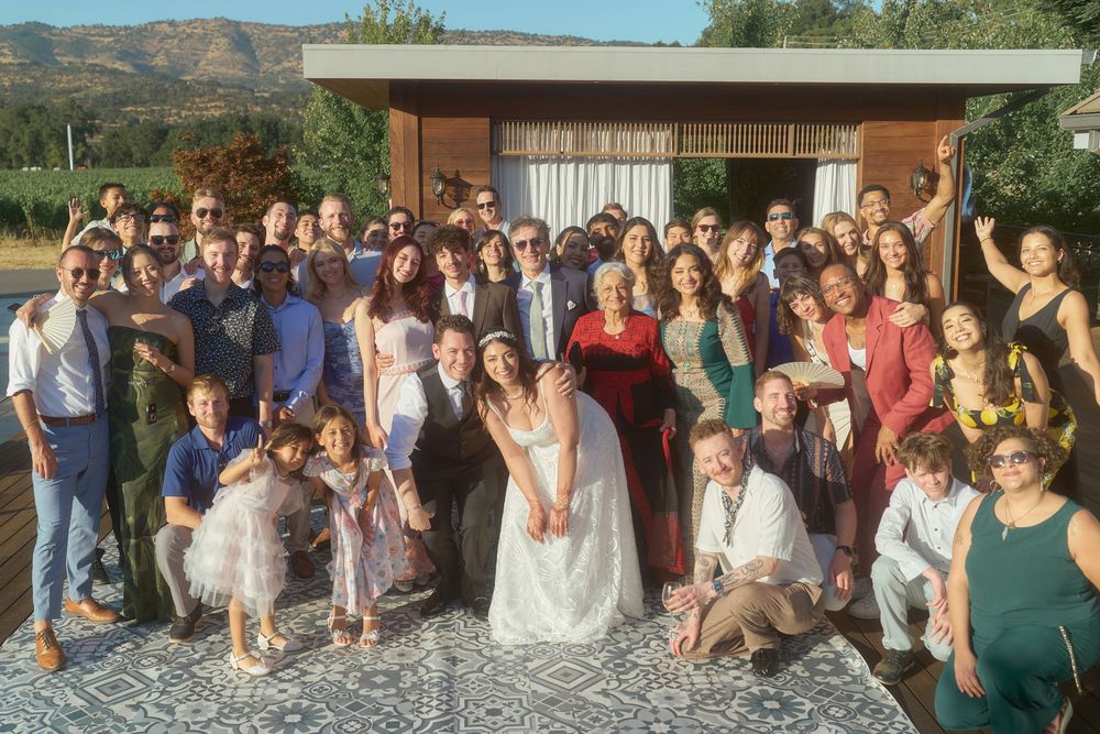 Large wedding party group photo with bride and groom surrounded by family and friends outdoors.