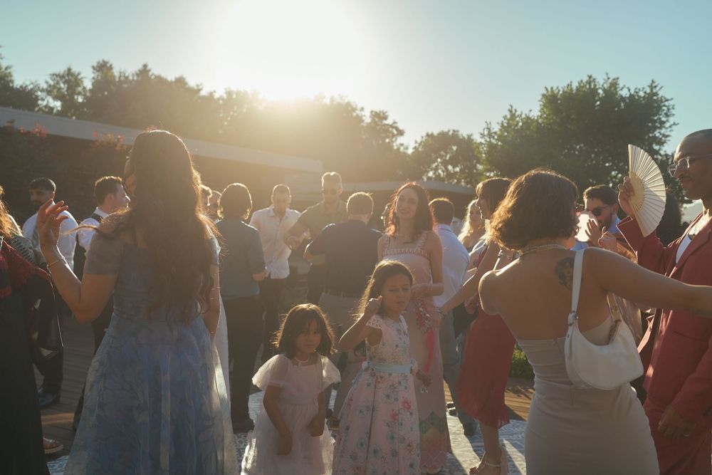Wedding guests dancing outdoors in golden sunset light with a young flower girl among the crowd.