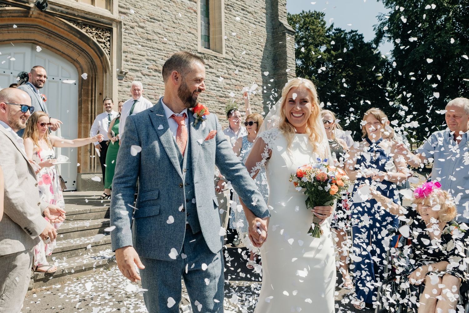 A couple stands and smiles together during an outdoor celebration with greenery in the background.