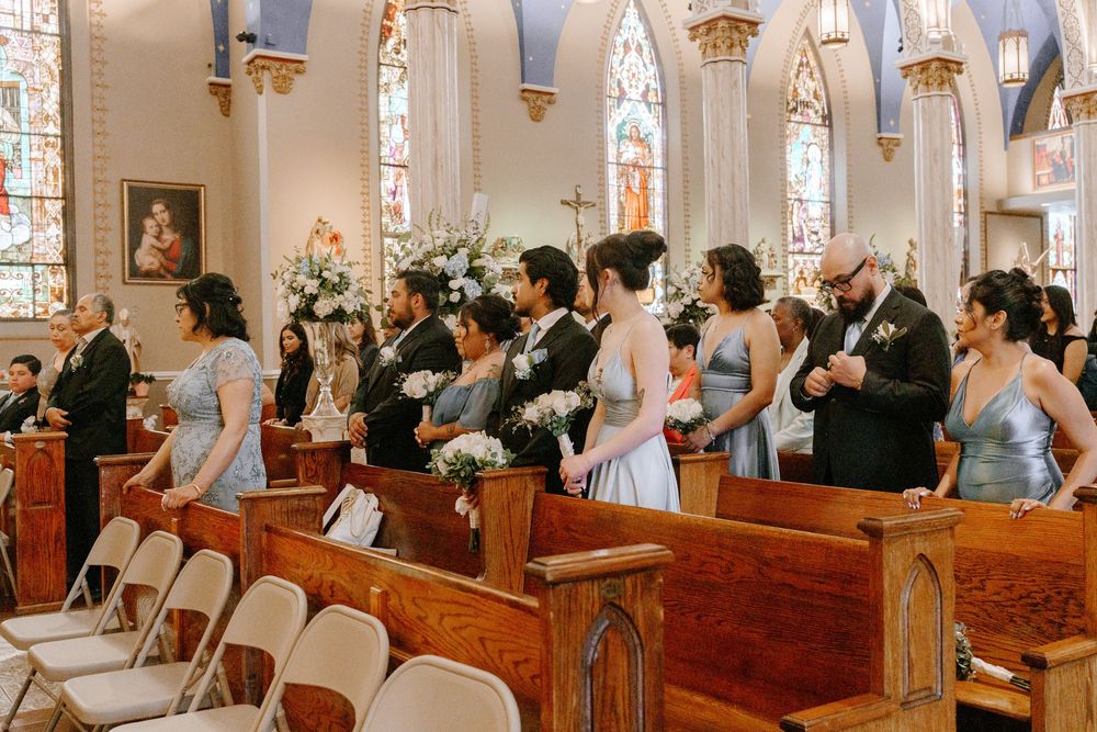 Wedding guests stand in a church pew during a ceremony, with stained glass windows and ornate architecture visible.