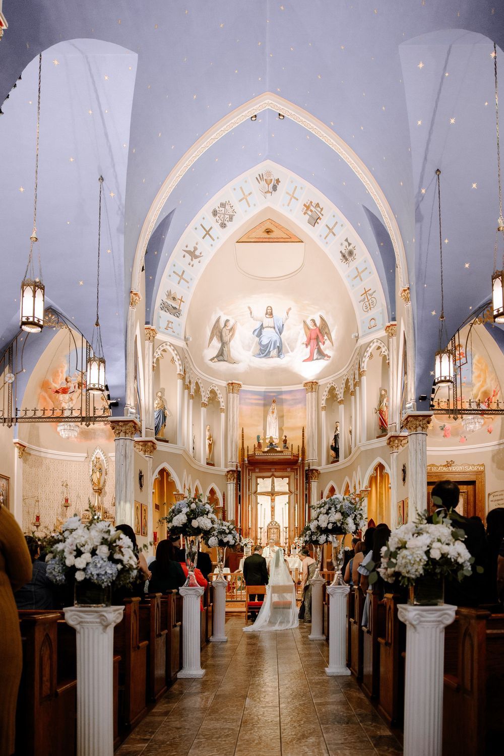 Interior view of a gothic-style church with white decorative arches, stained glass windows and floral arrangements on columns.