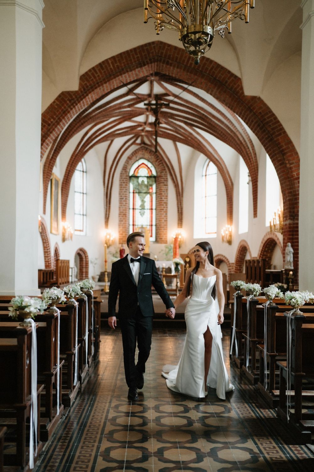 Couple walking through a grand church hallway with gothic arches and ornate chandeliers.