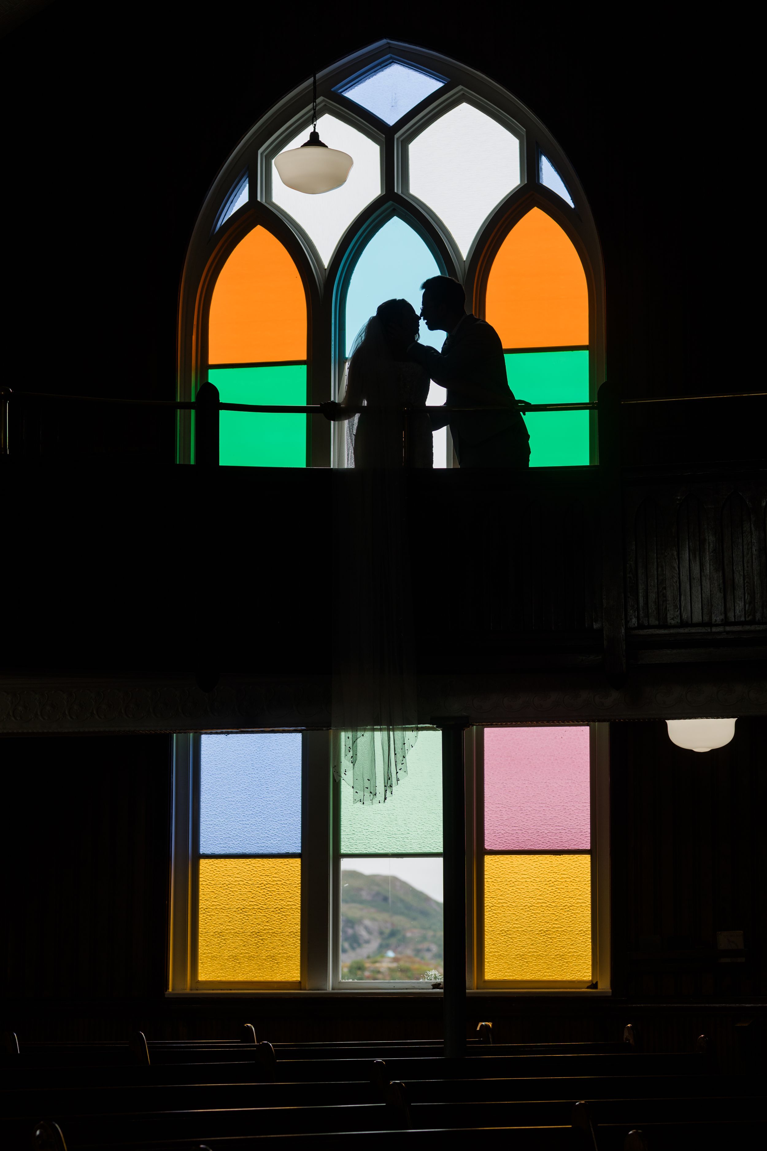 Silhouetted figures stand in a church window with colorful stained glass panels featuring orange, green, and blue hues.