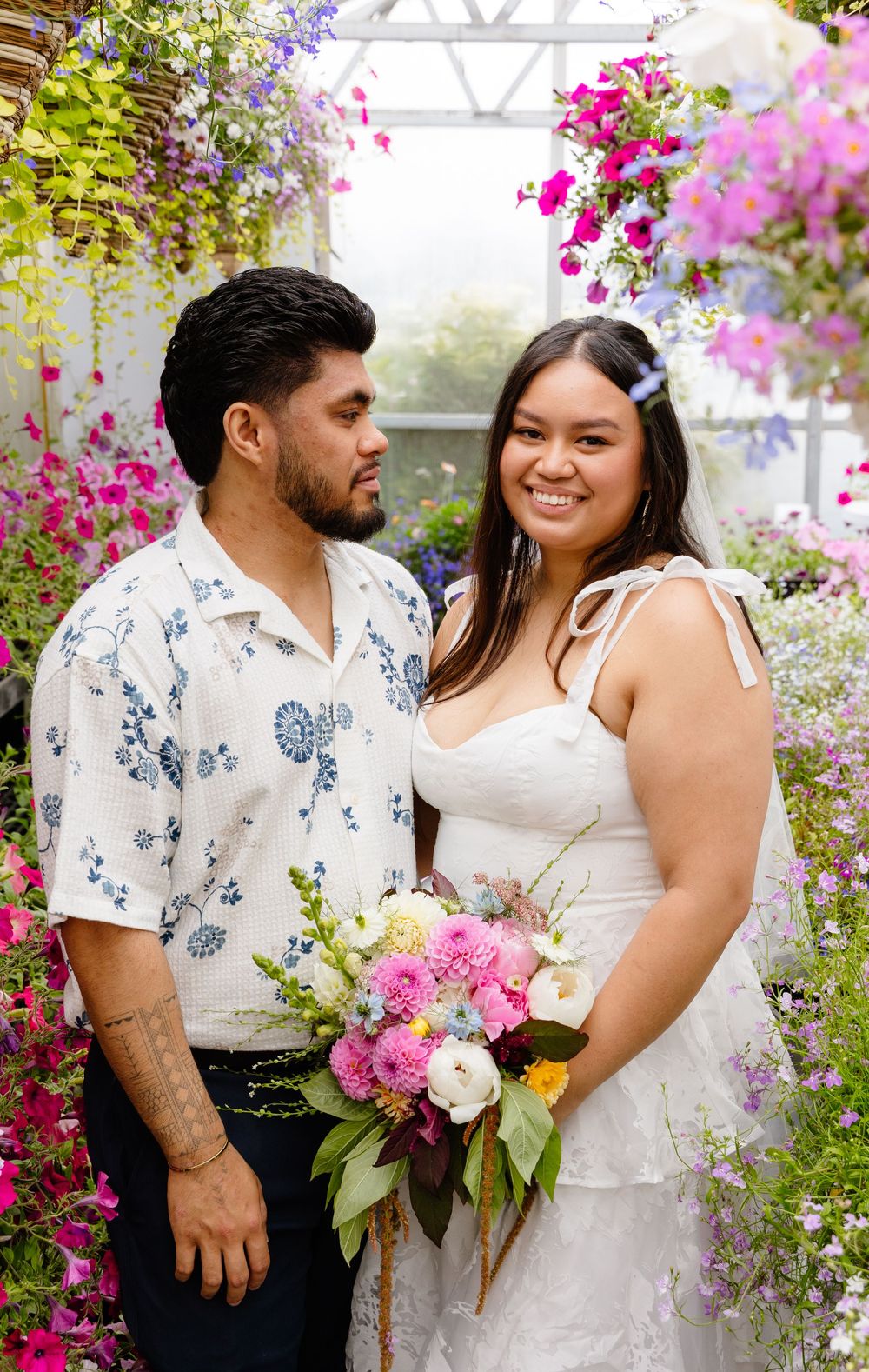 A couple poses in a garden greenhouse surrounded by vibrant pink and purple hanging flowers.
