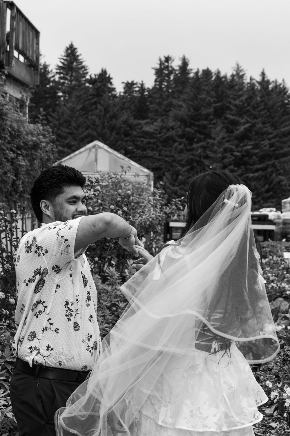 A couple during a wedding photoshoot in a garden, with the bride's veil flowing in the wind in black and white.