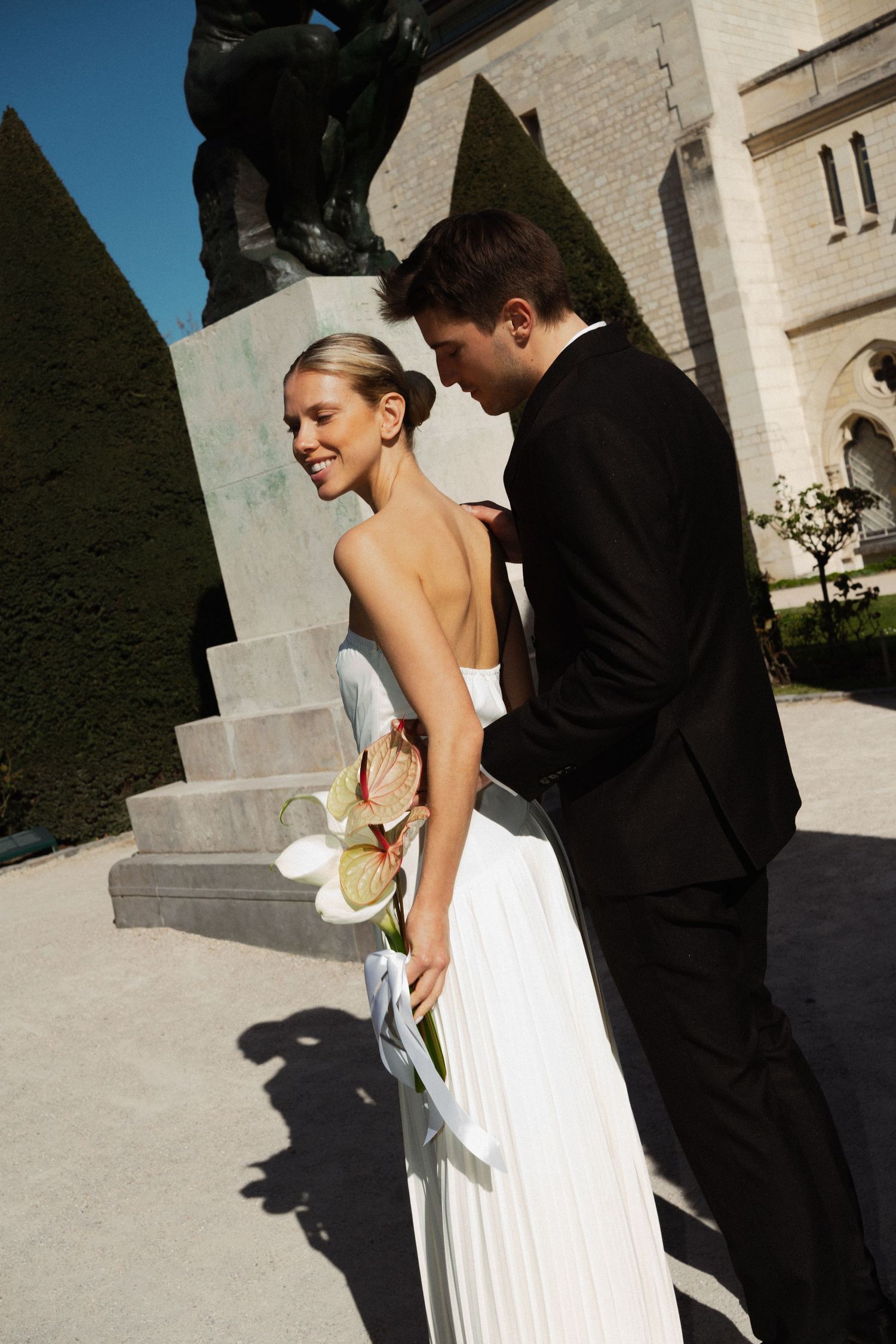 A romantic wedding moment between a couple in formal attire standing next to a stone wall in bright sunlight.