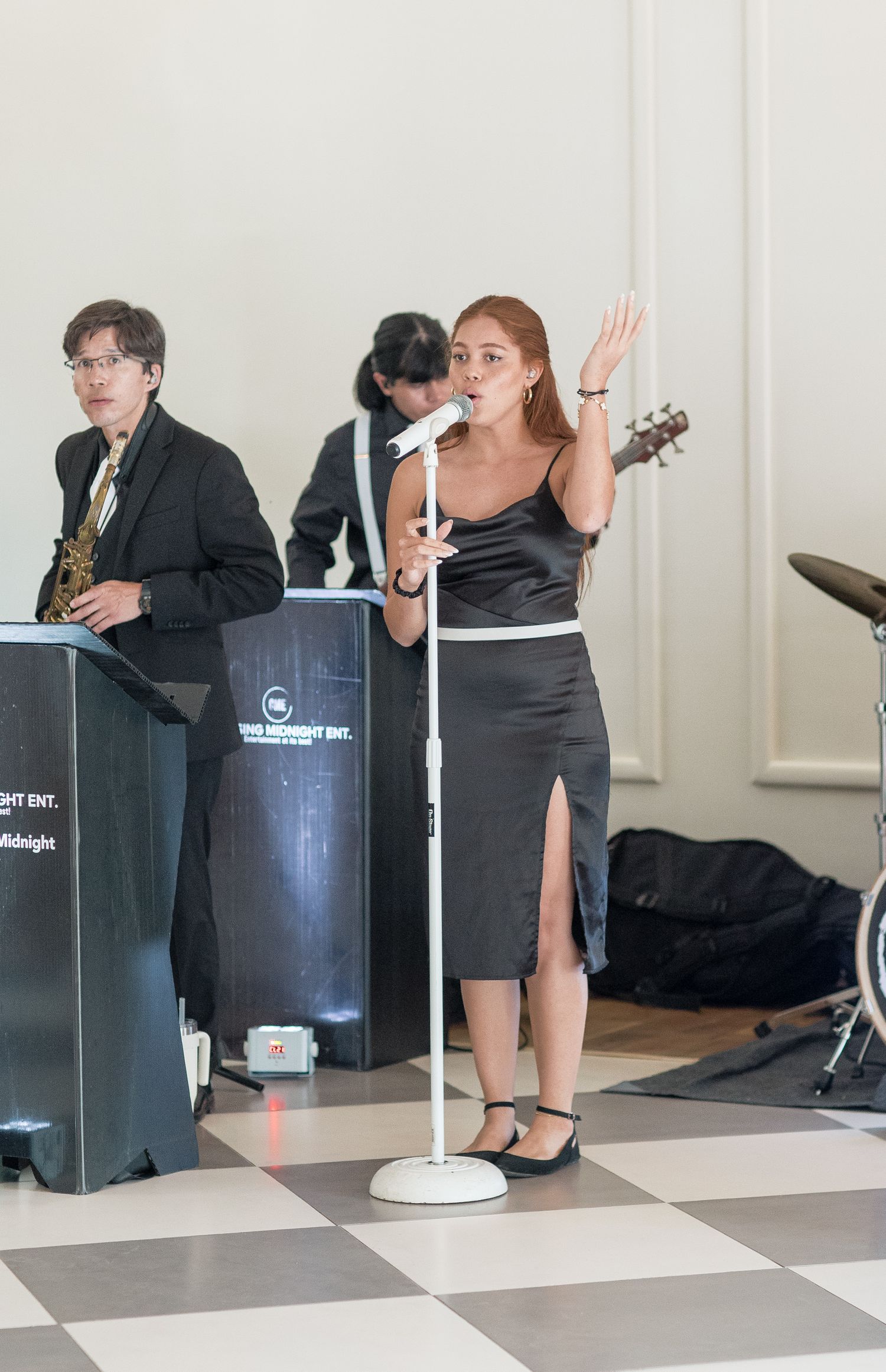Speakers and musicians prepare on a stage with black equipment and white cables against a light colored wall.