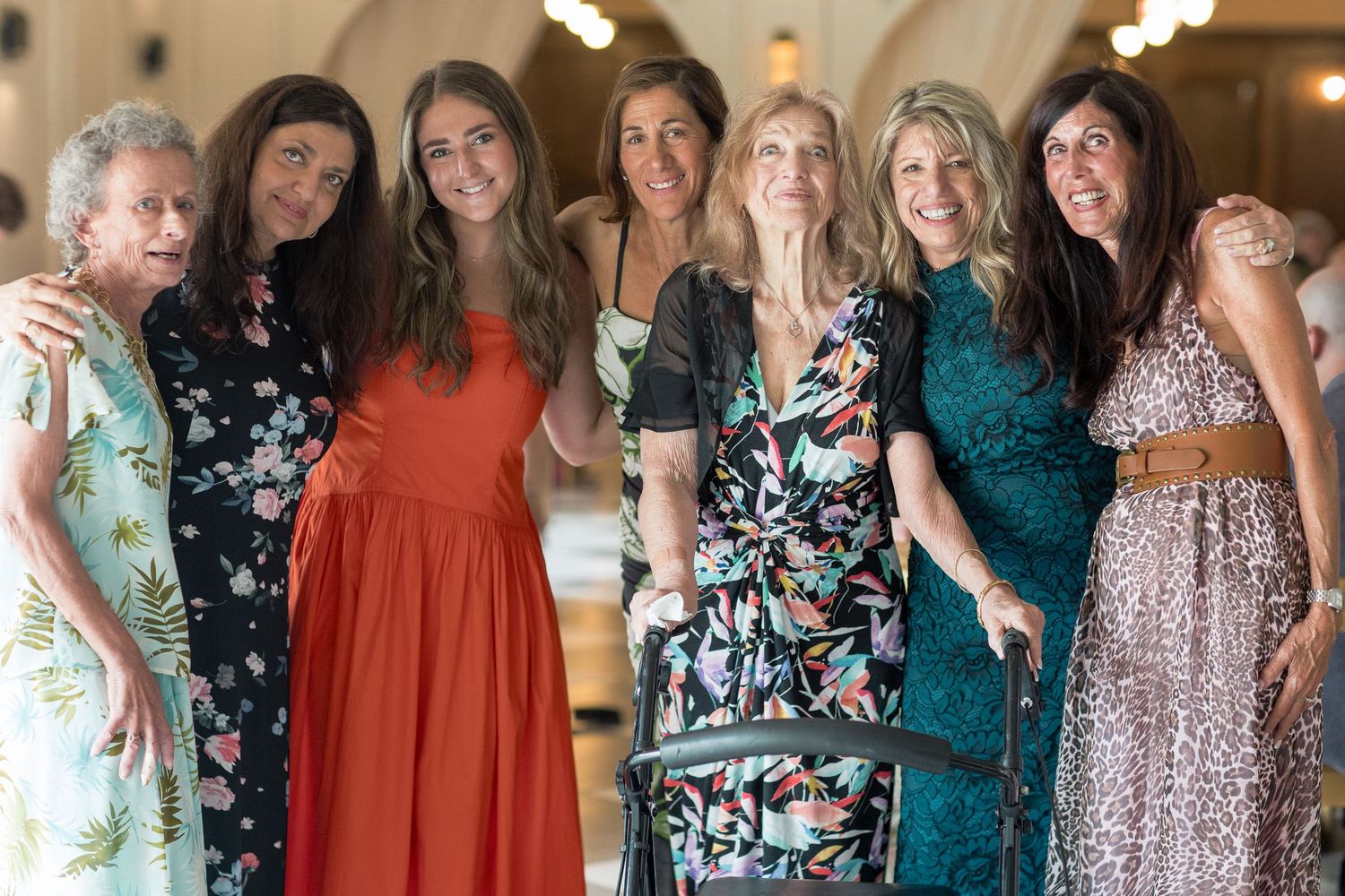 A group of women wearing colorful dresses and formal attire stand together posing at an elegant indoor event.