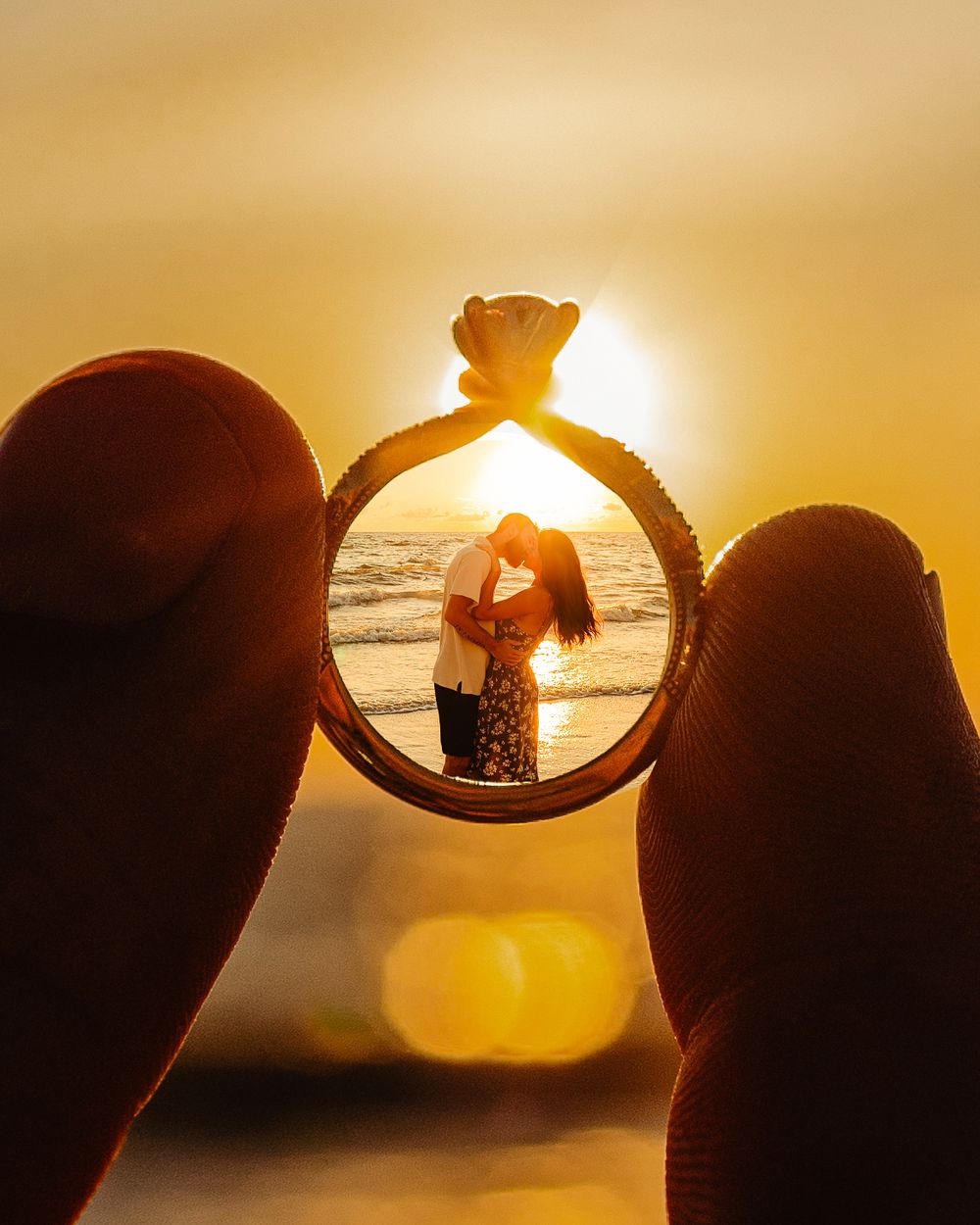Two silhouettes hold engagement ring at sunset, framing a romantic beach scene perfectly through the ring's diamond.