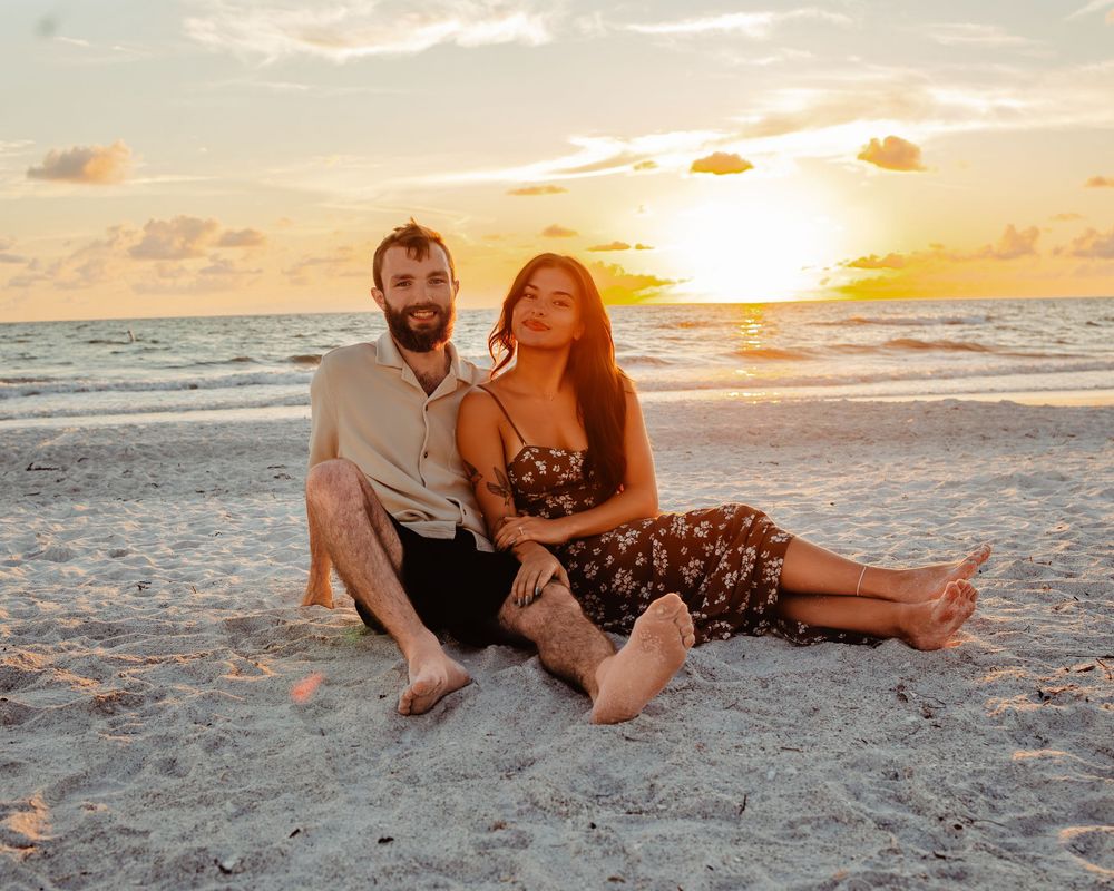 A couple sits together on a sandy beach during golden sunset against a vibrant orange and blue sky.