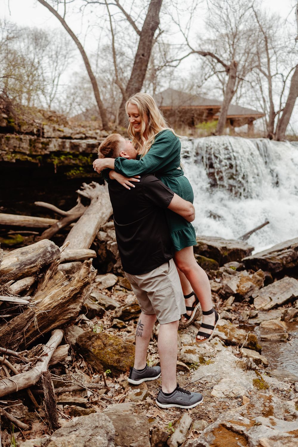 A couple embraces near a scenic waterfall on rocky terrain surrounded by bare trees on an autumn day.
