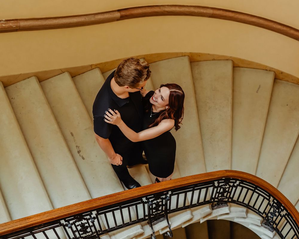 A couple in black outfits embrace on an elegant spiral staircase with cream-colored steps and dark wooden railing.