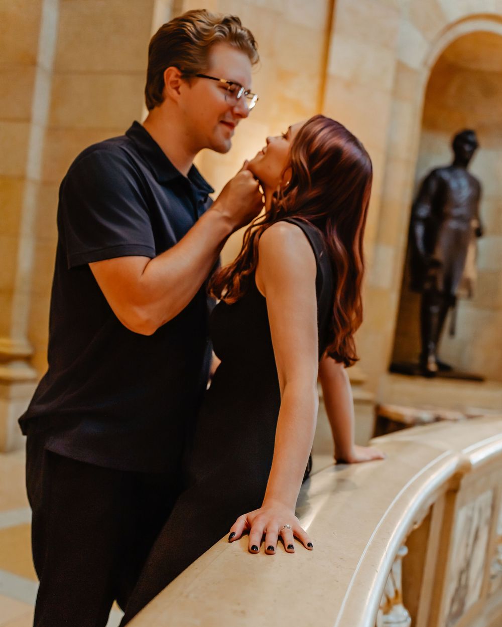 A romantic couple in black attire share an intimate moment in front of arched stone columns with a statue silhouette nearby.