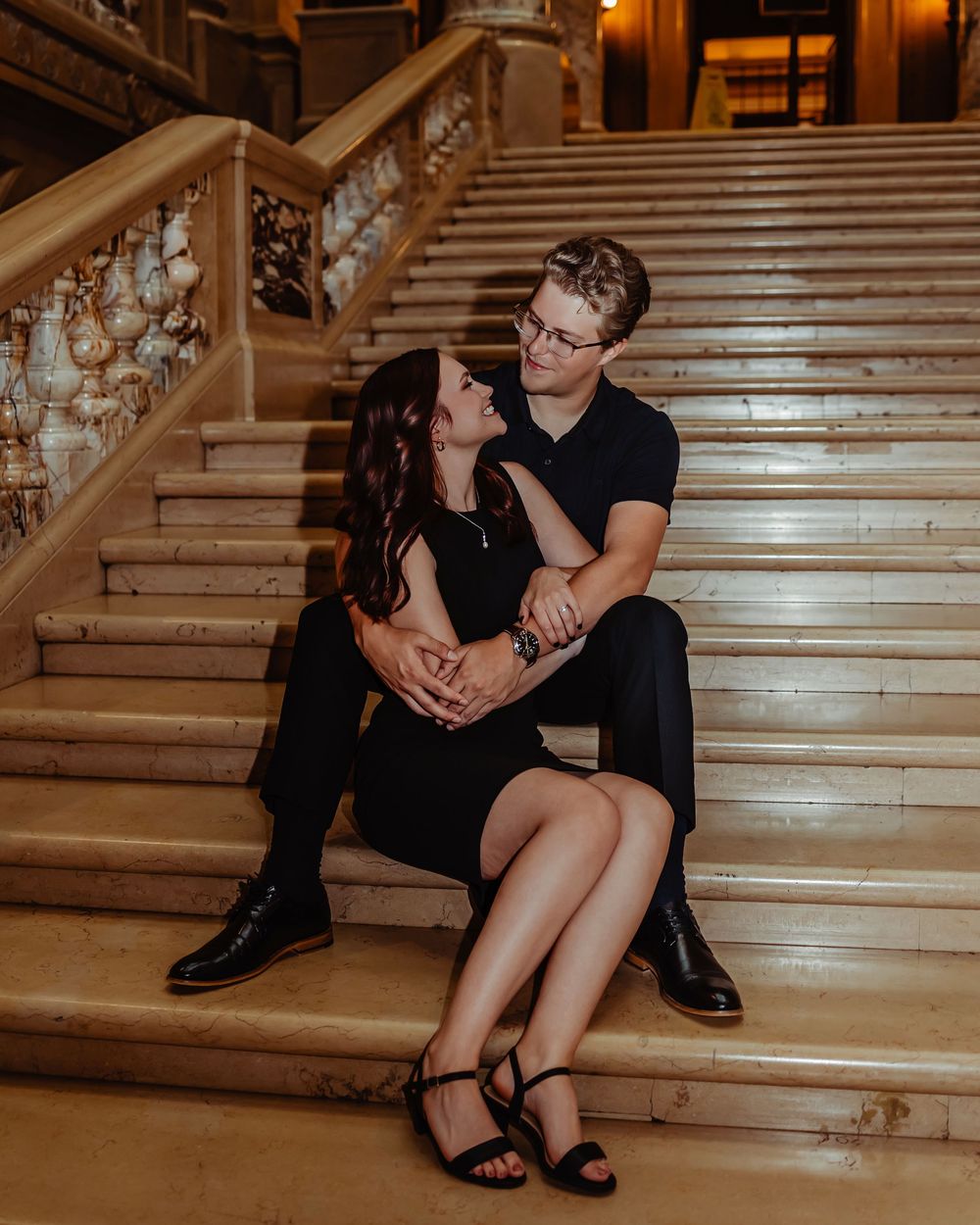 A romantic couple sits together on an elegant marble staircase in a warmly lit interior setting.
