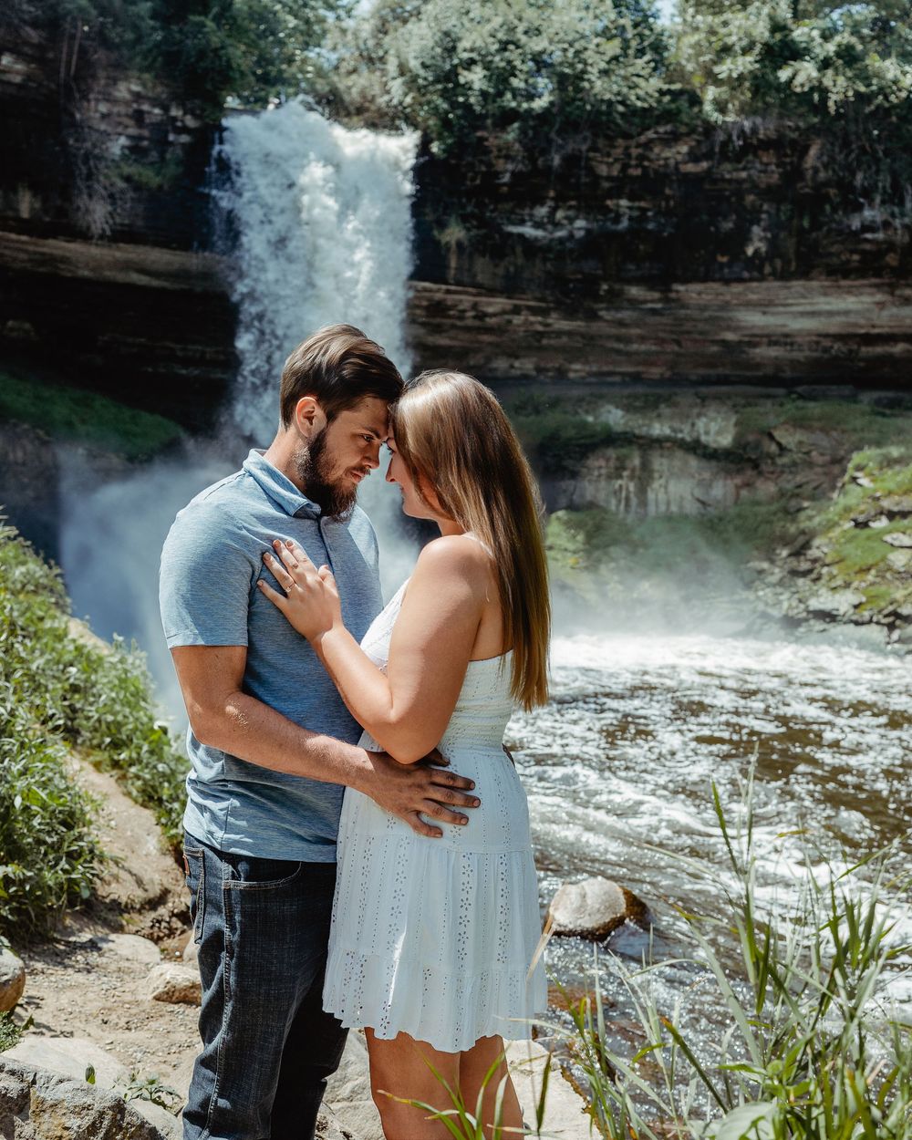 A romantic couple embraces in front of a majestic waterfall cascading down rocky cliffs on a sunny day.
