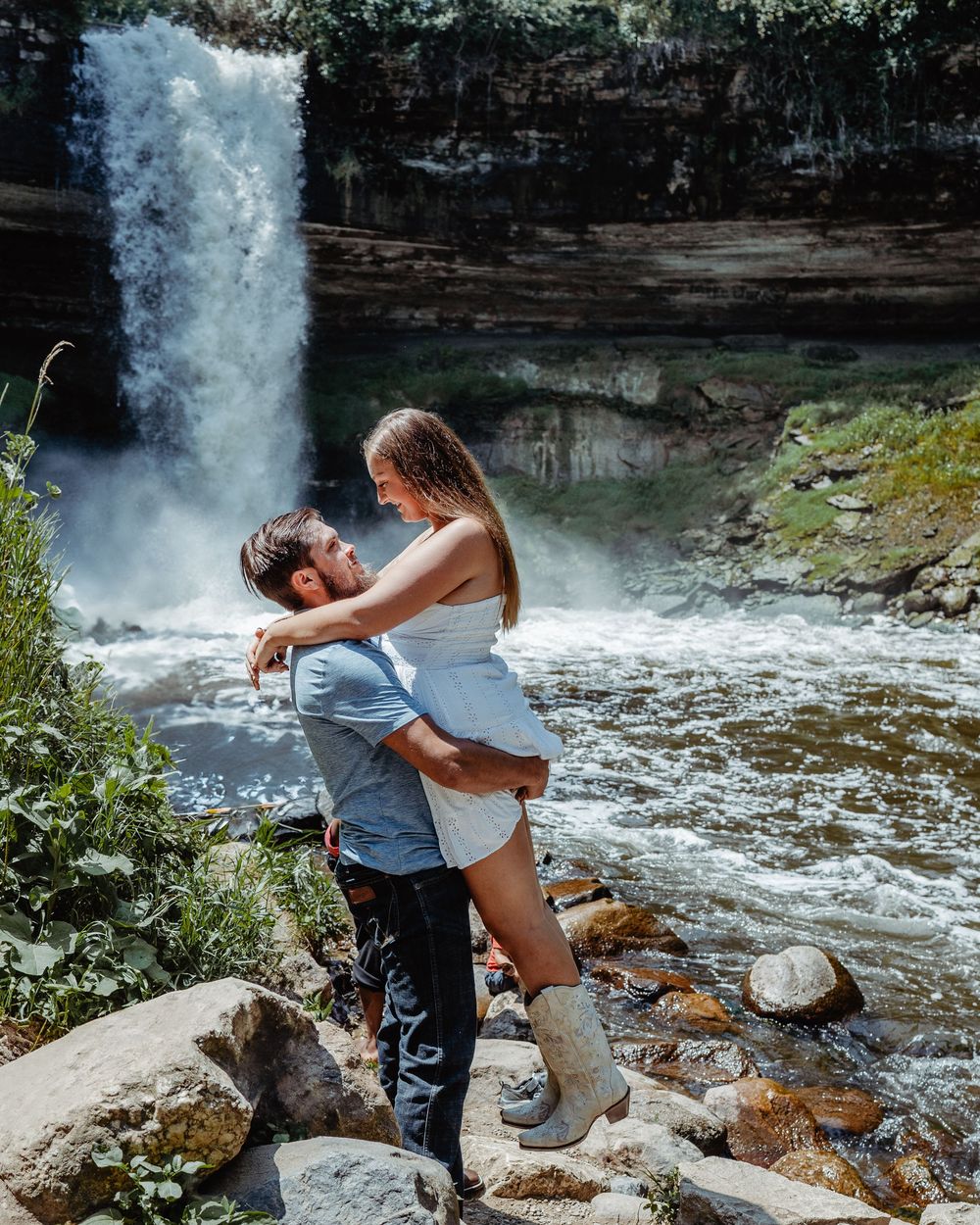 A couple embraces romantically by a cascading waterfall on rocky terrain in a scenic natural setting.