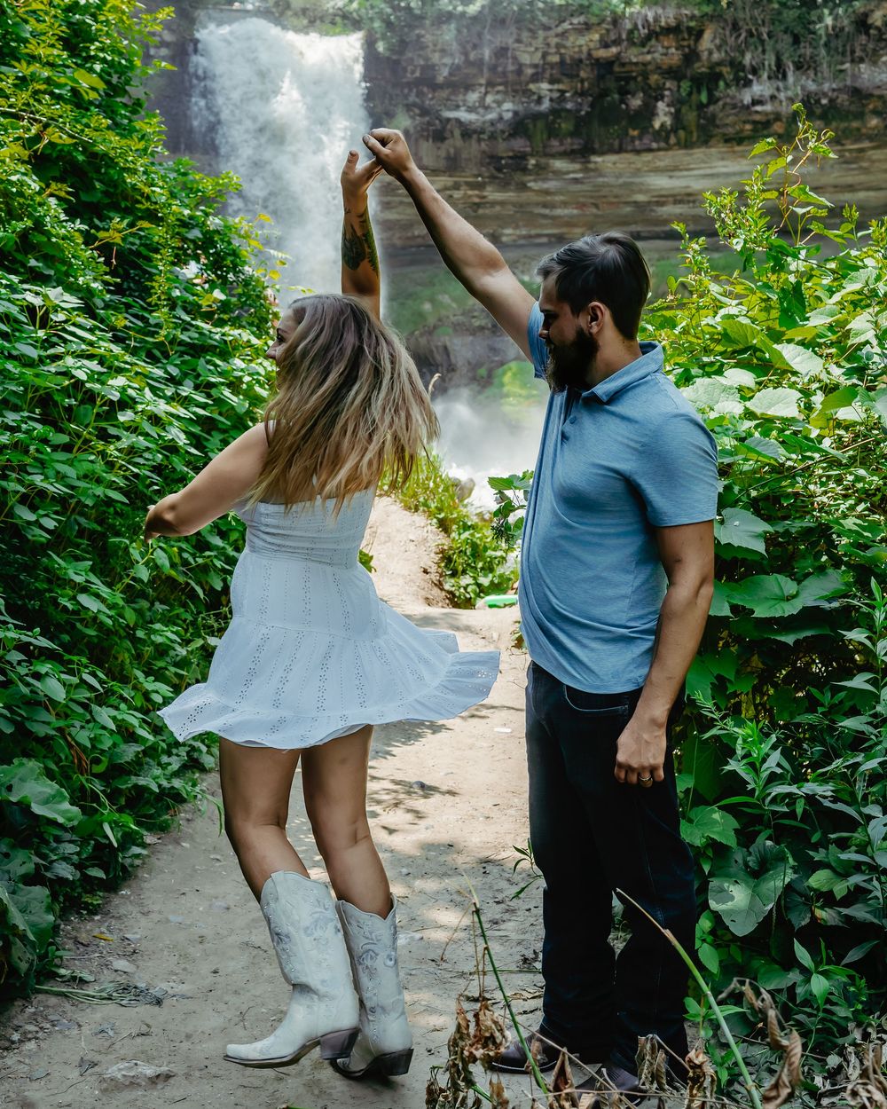 A couple dances together on a scenic trail with a majestic waterfall backdrop at a state park.