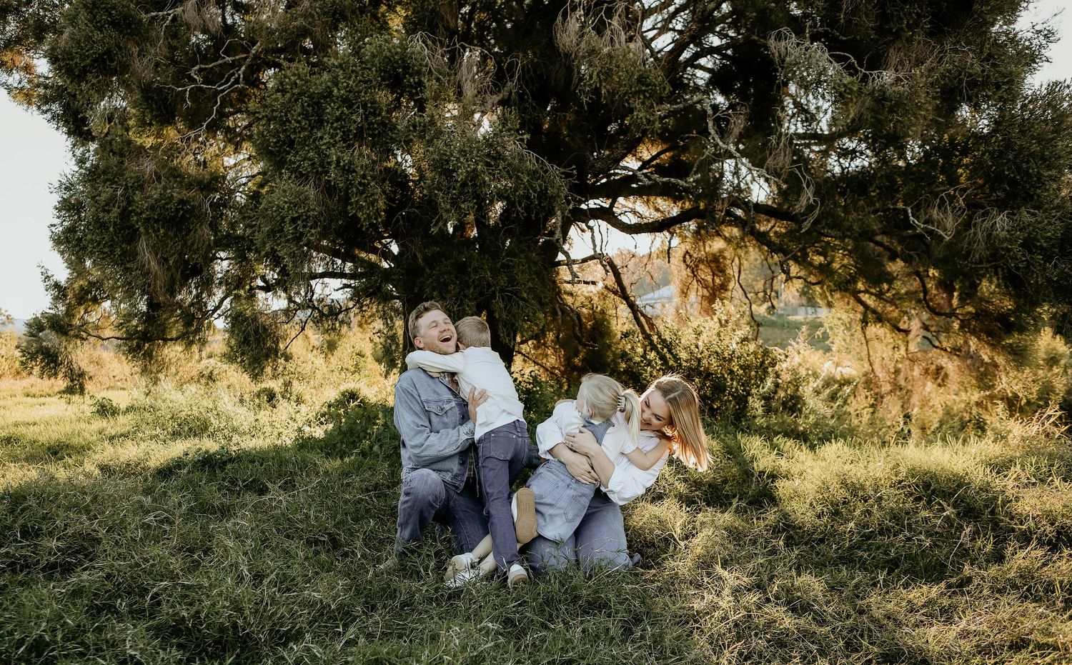 Family photo session in a rustic outdoor setting with golden sunlight filtering through trees during sunset.