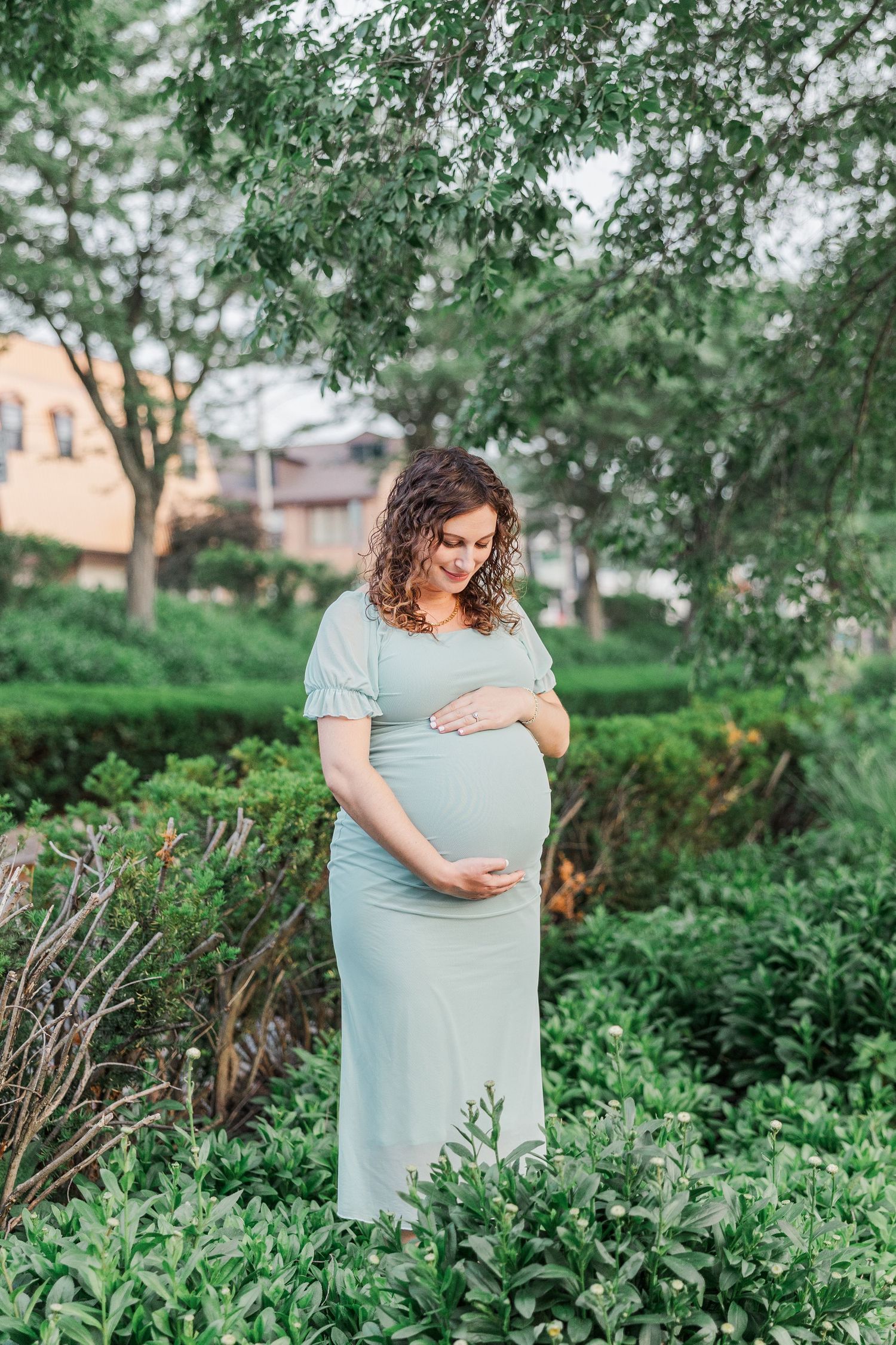 Expectant mother in a mint green dress poses in a lush garden setting during maternity photoshoot.