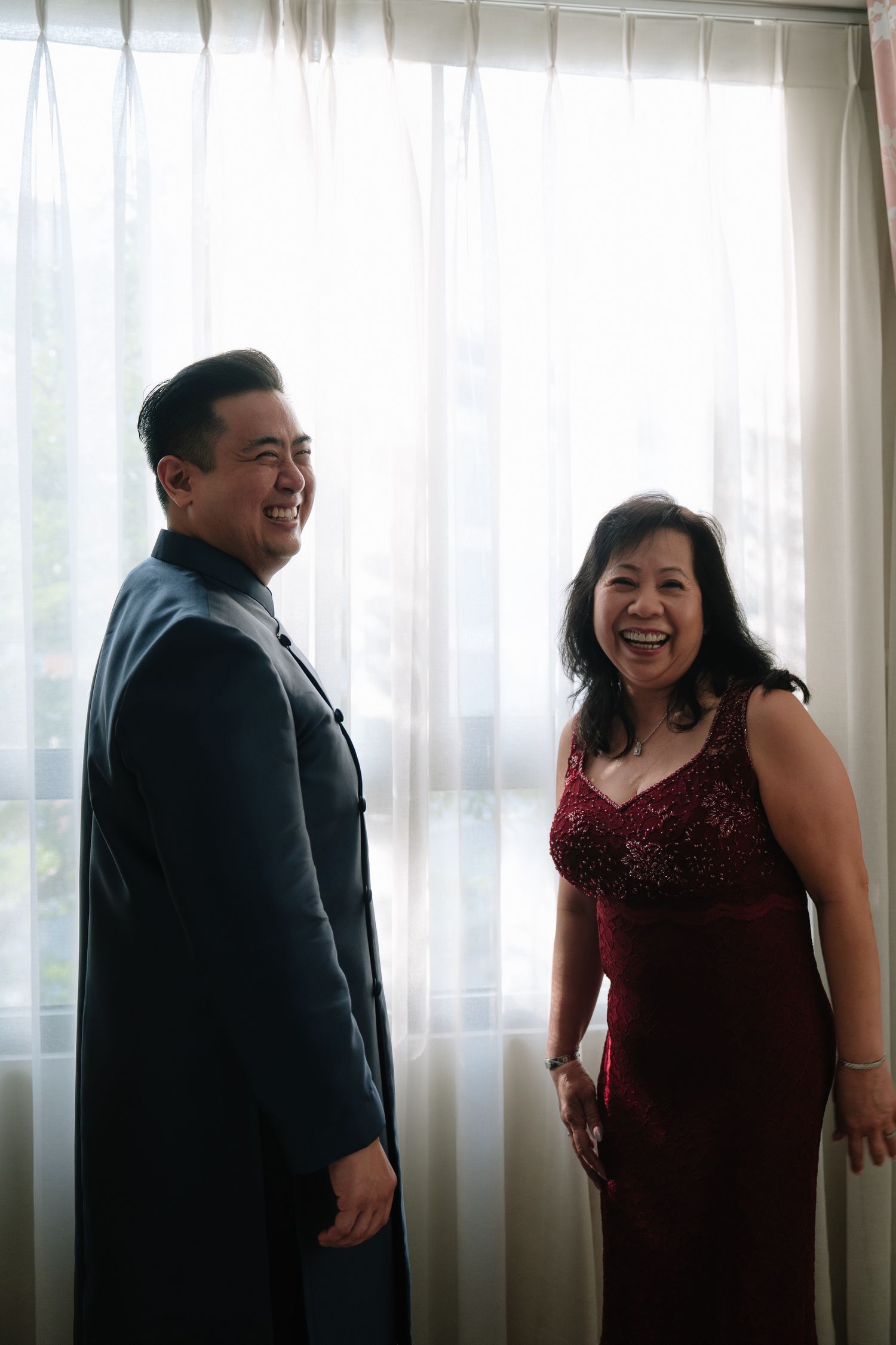 Groom in blue mandarin suit laughing with woman in red dress by window.