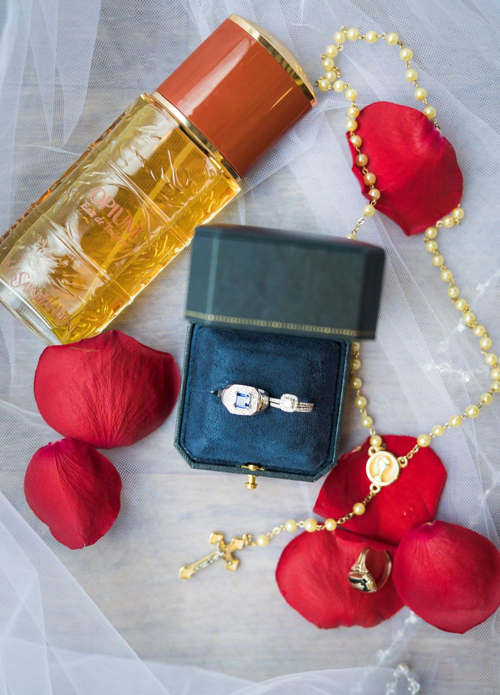 Wedding details flatlay with perfume bottle, engagement ring box, pearl necklace and red rose petals on white tulle.