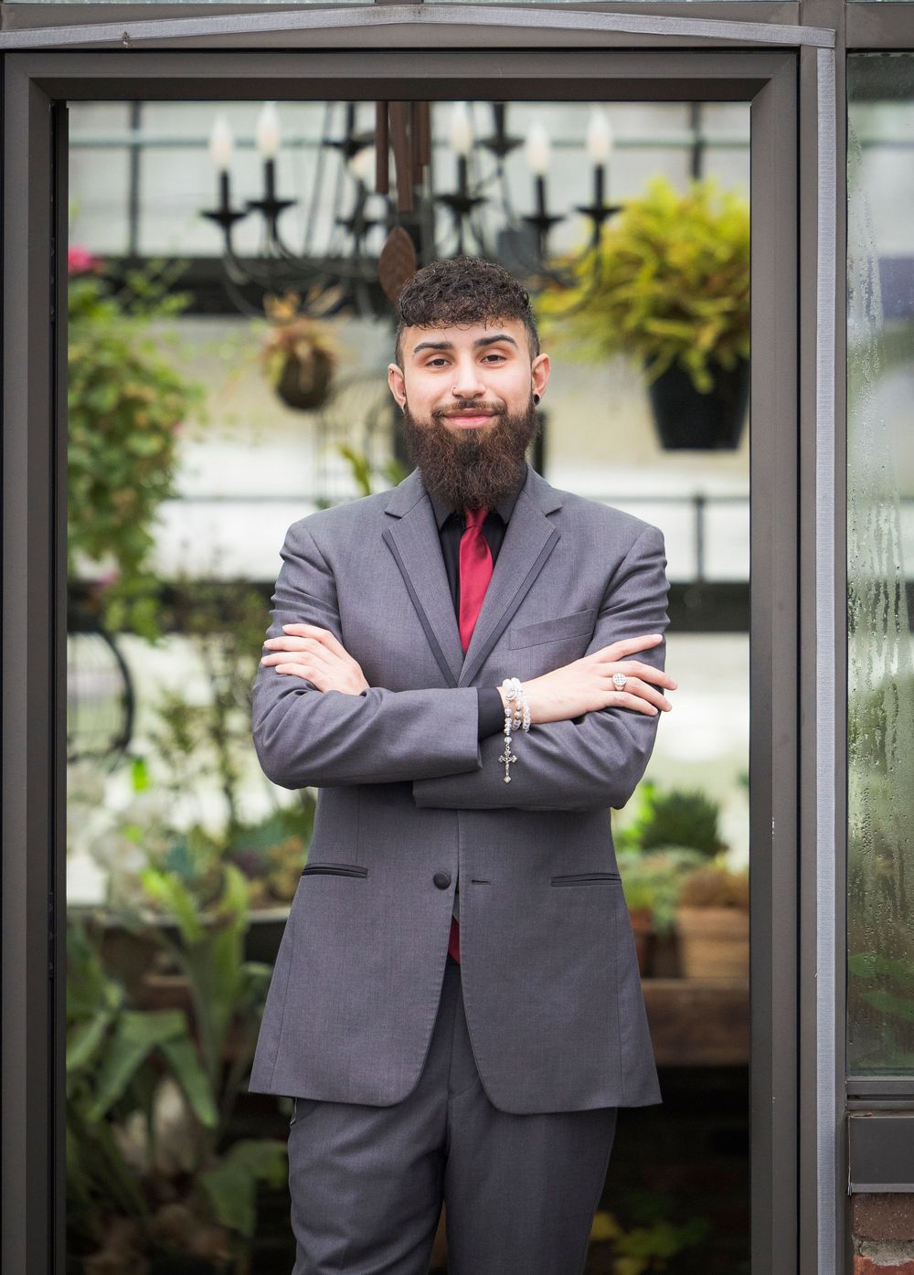 Professional wedding portrait of a groom in a grey suit and red tie standing against a glass wall with greenery.