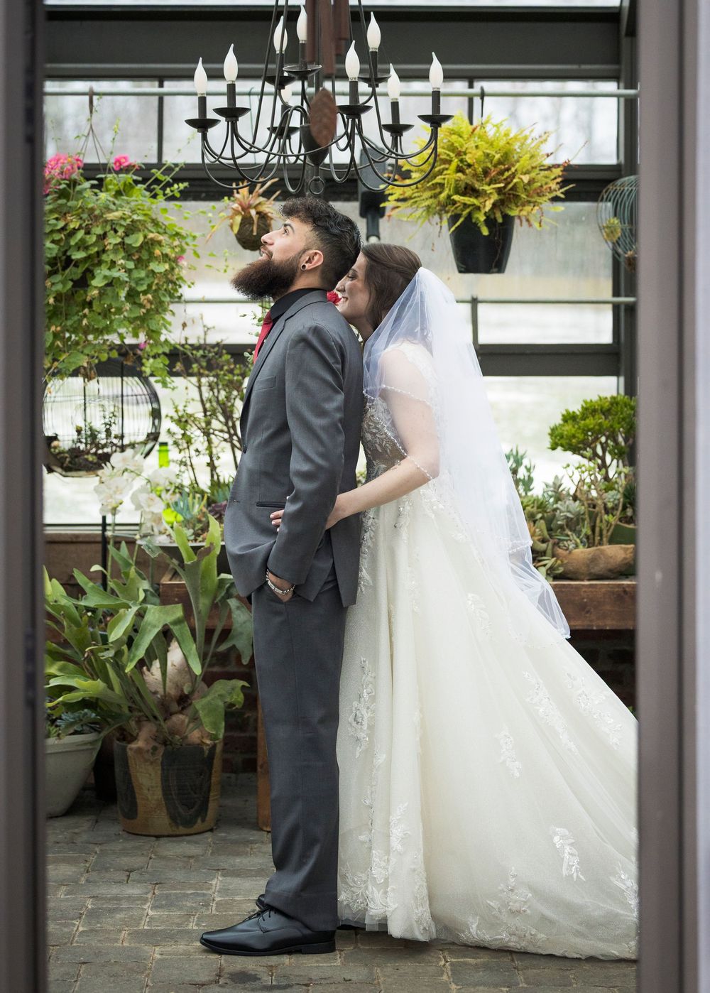 A couple poses in a greenhouse setting with lush plants and a black chandelier overhead.