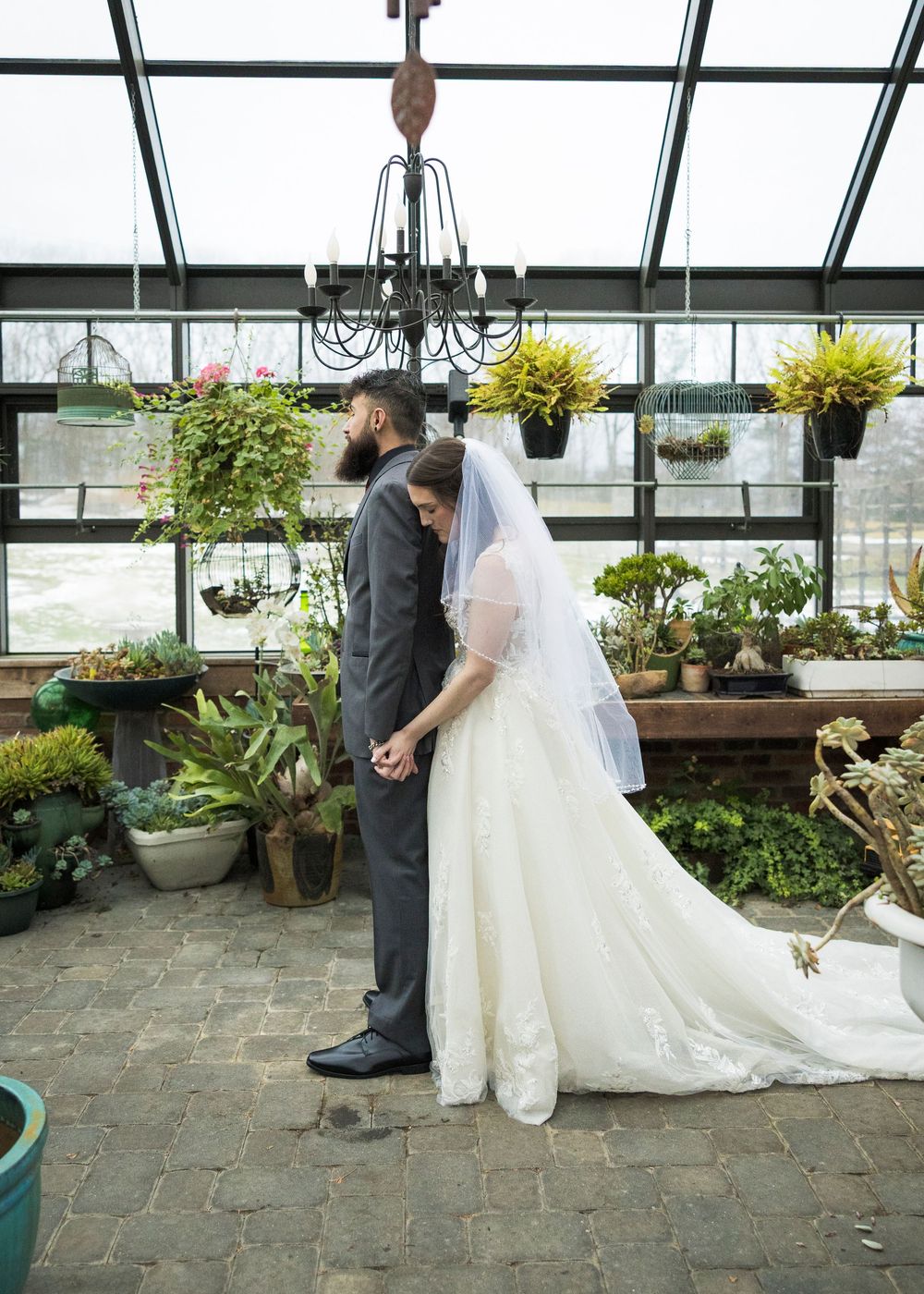 Wedding couple poses in a greenhouse setting with hanging plants and stone pavers.