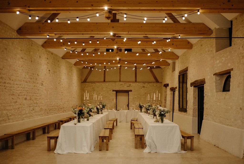 Rustic barn wedding reception hall with string lights, long white tables, and wooden benches.