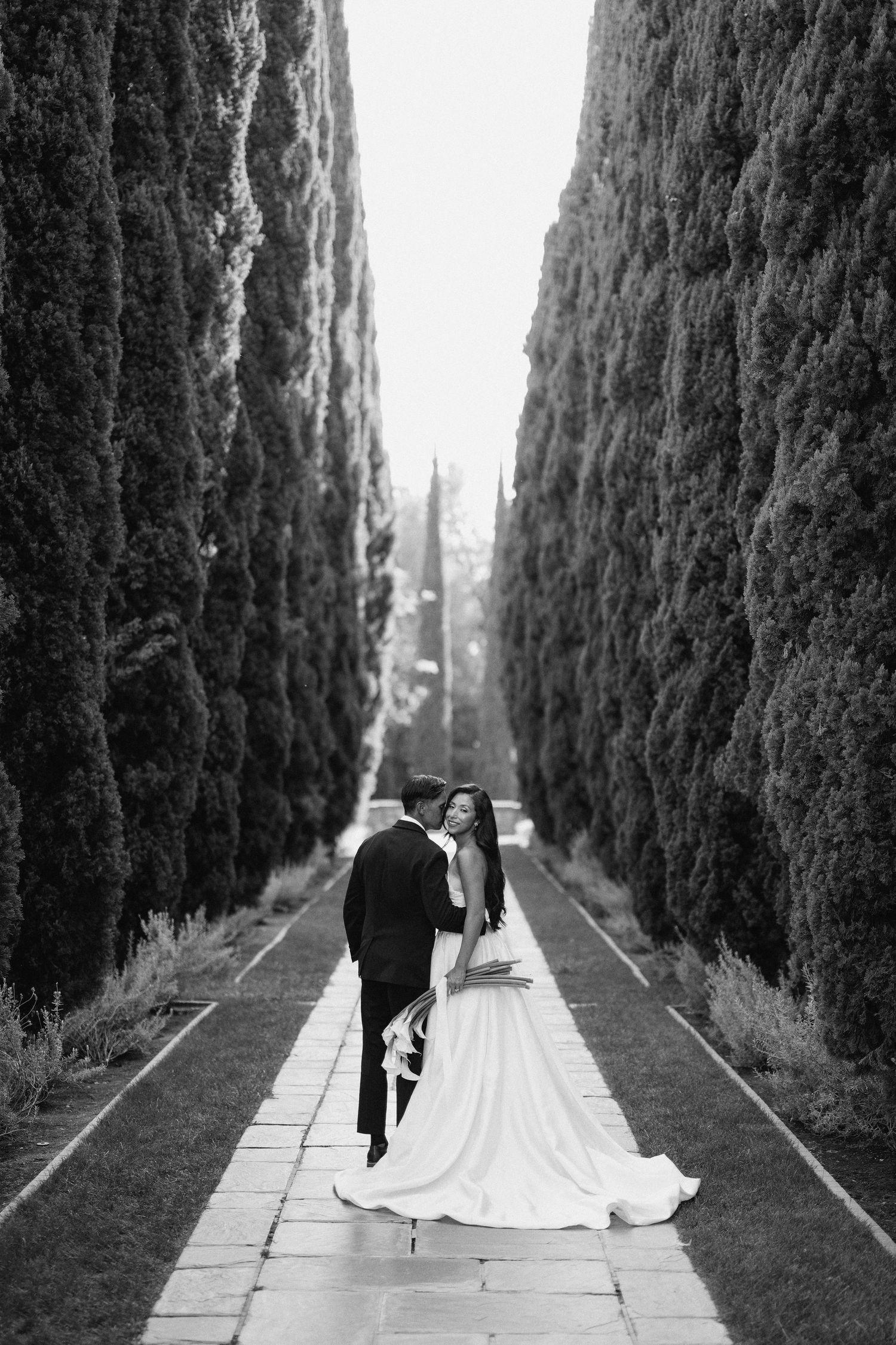 A romantic black and white series of four photos showing a couple walking down a cypress tree-lined path.