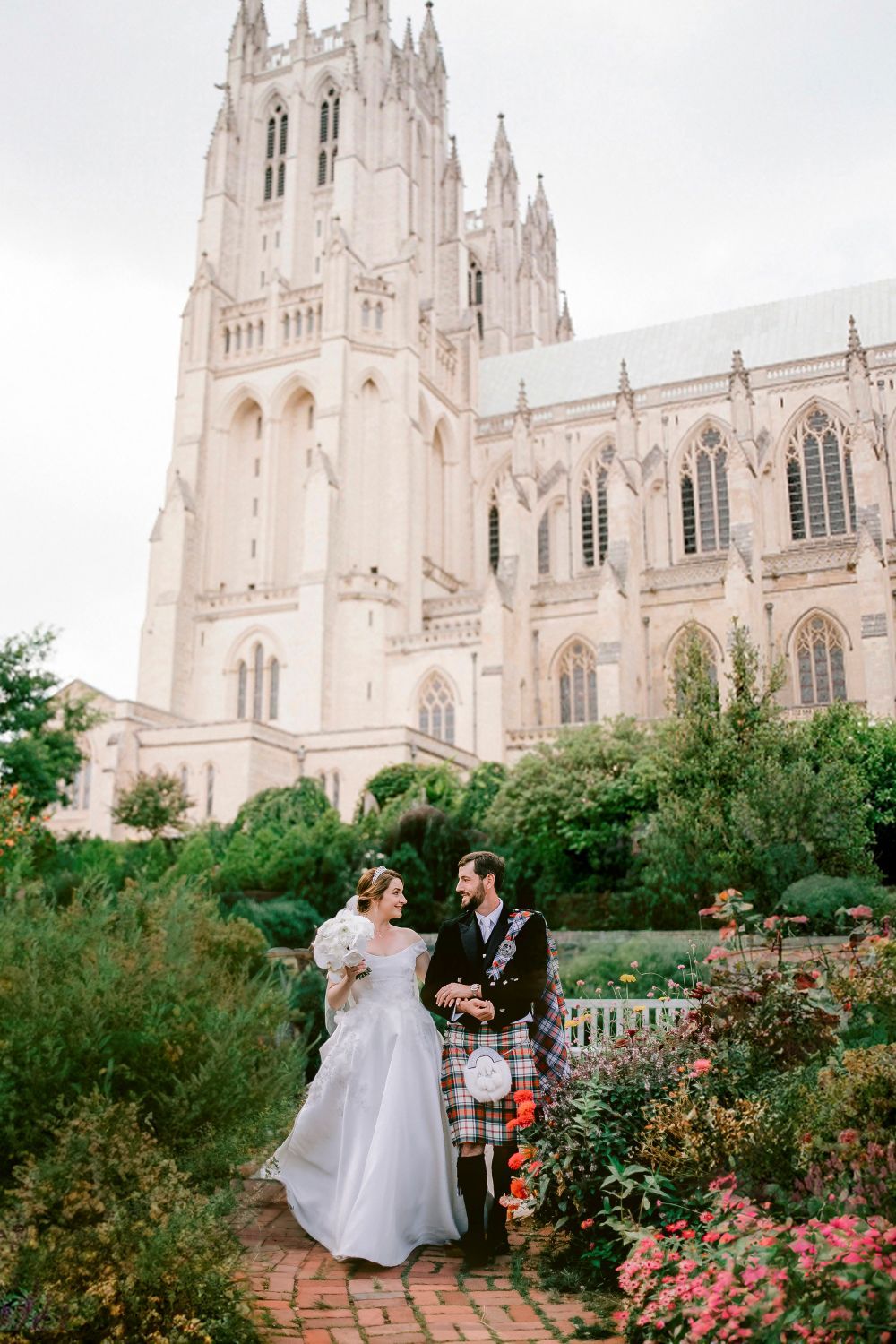 A couple in wedding attire poses in front of a grand Gothic cathedral with ornate spires and beautiful gardens.