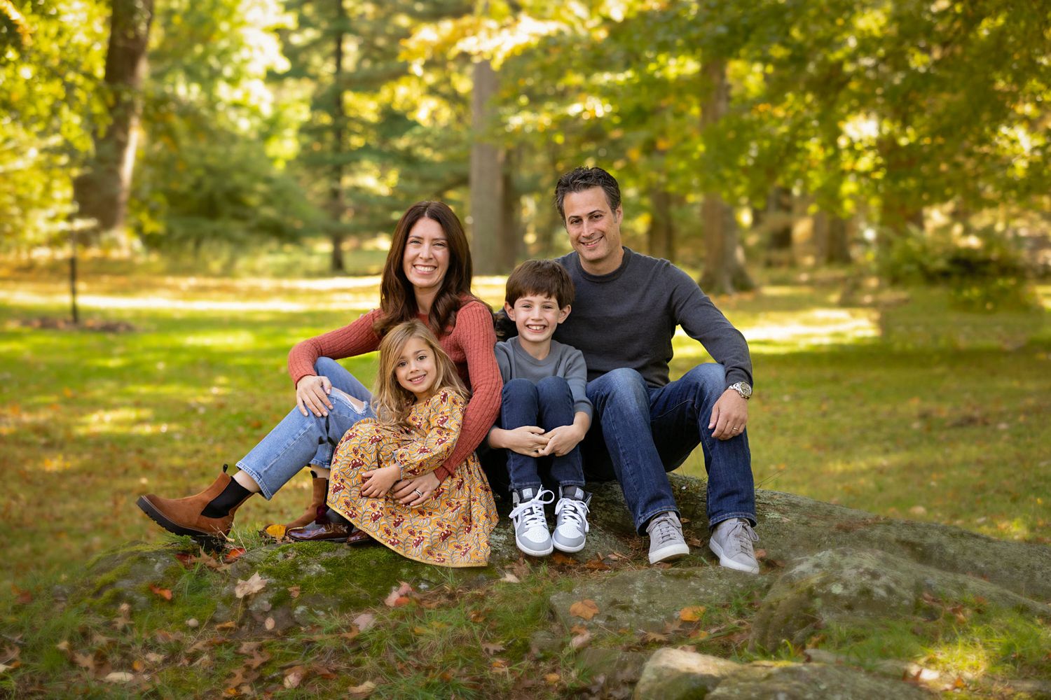 A family sits together outdoors on a sunny autumn day in a park with golden leaves in the background.
