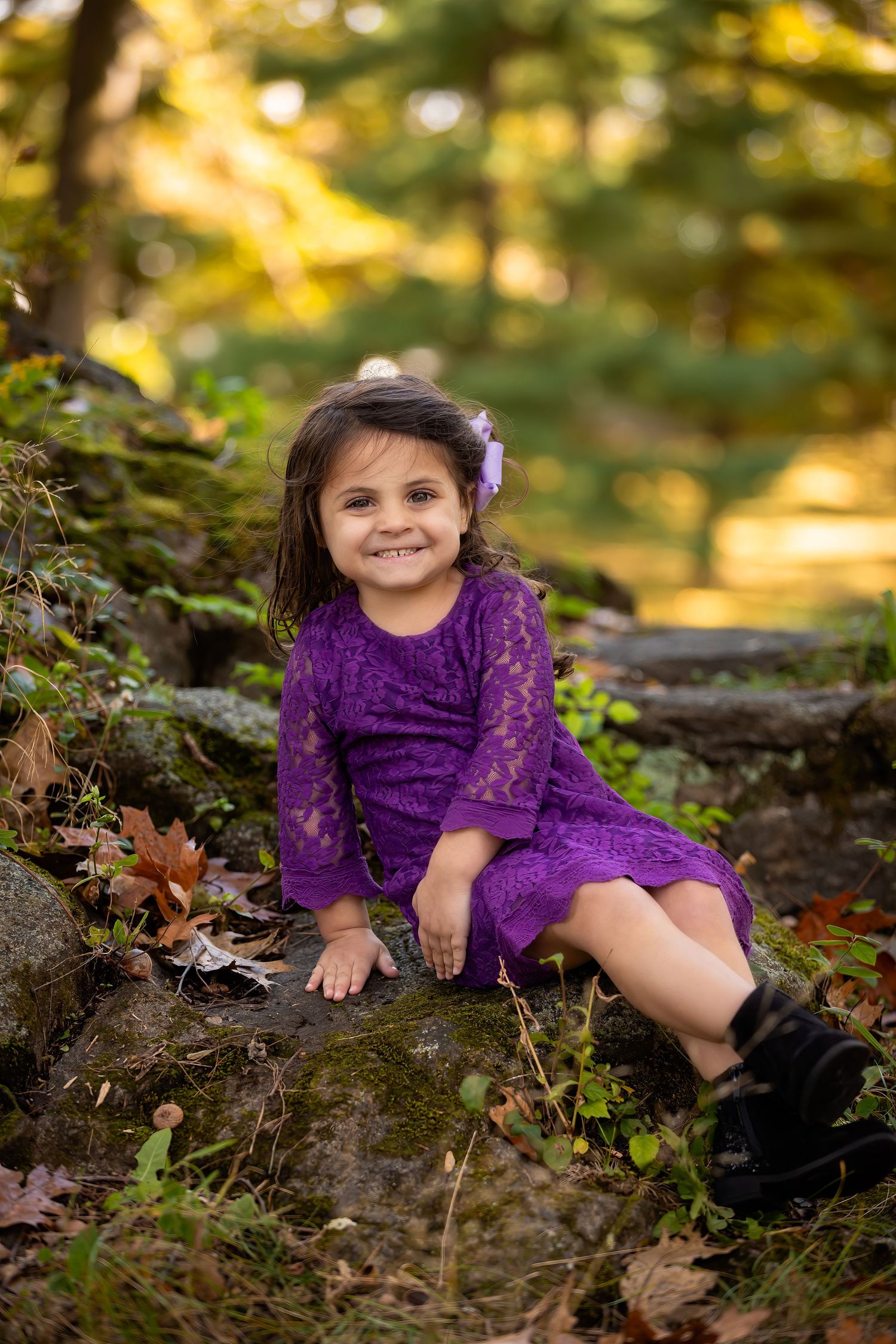 A child in a purple dress lies on autumn leaves and moss-covered rocks in a sunlit outdoor setting.