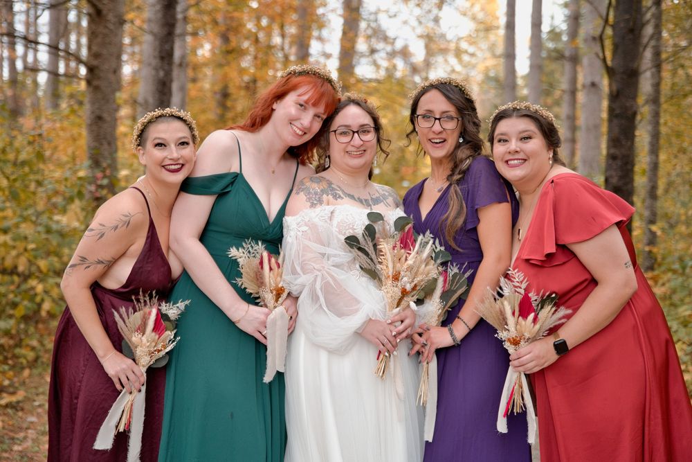 Smiling group dressed in jewel-toned dresses share joyful embraces during autumn wedding photoshoot in wooded setting.