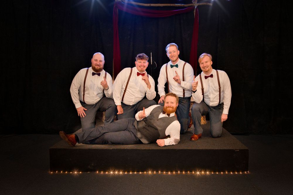 Five performers in matching suspenders and dress shirts pose together on a dark stage with dramatic lighting and red curtains.