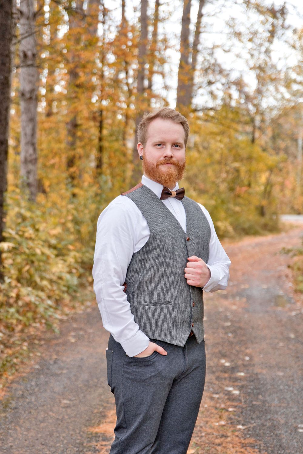 A groom in a grey vest and white shirt poses outdoors among autumn trees with golden foliage.