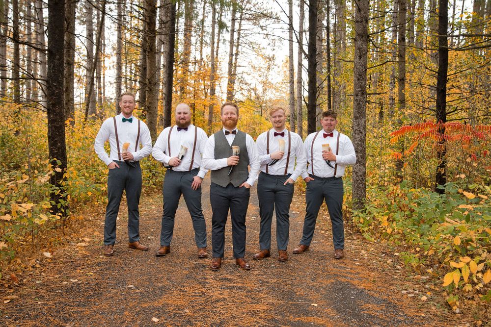 Groomsmen in matching attire stand together on a forest path surrounded by fall foliage.