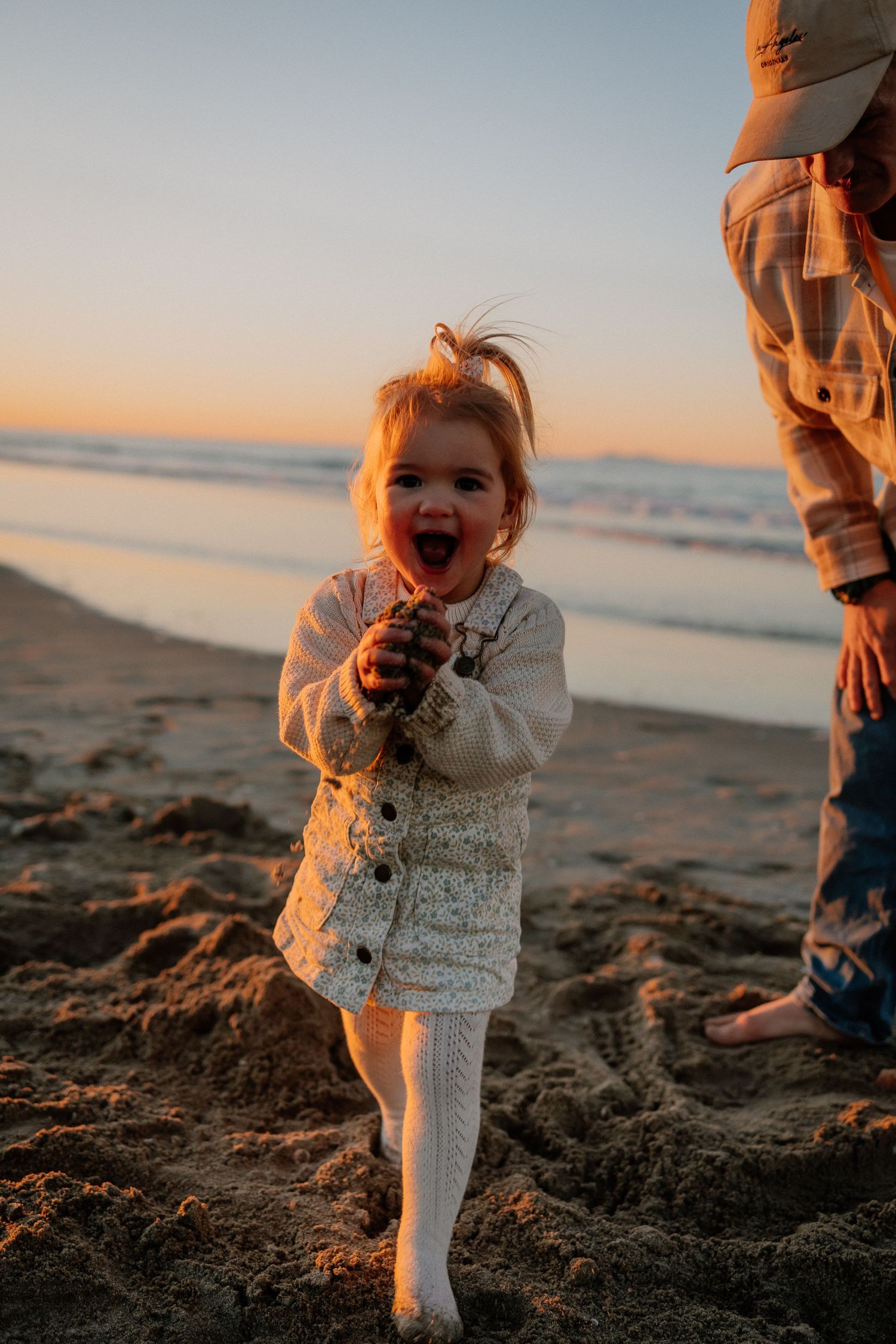 A small child in white clothing laughs joyfully on a beach at sunset while standing next to someone in casual wear.