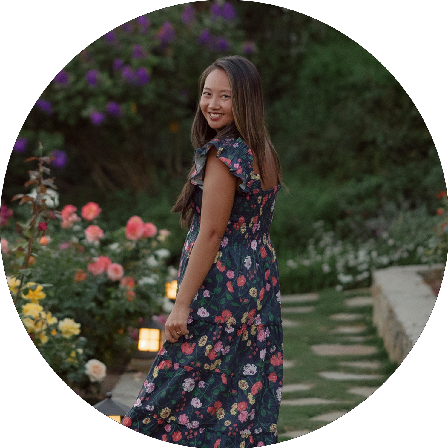 A woman in a floral dress smiles while standing in a garden with blooming flowers.