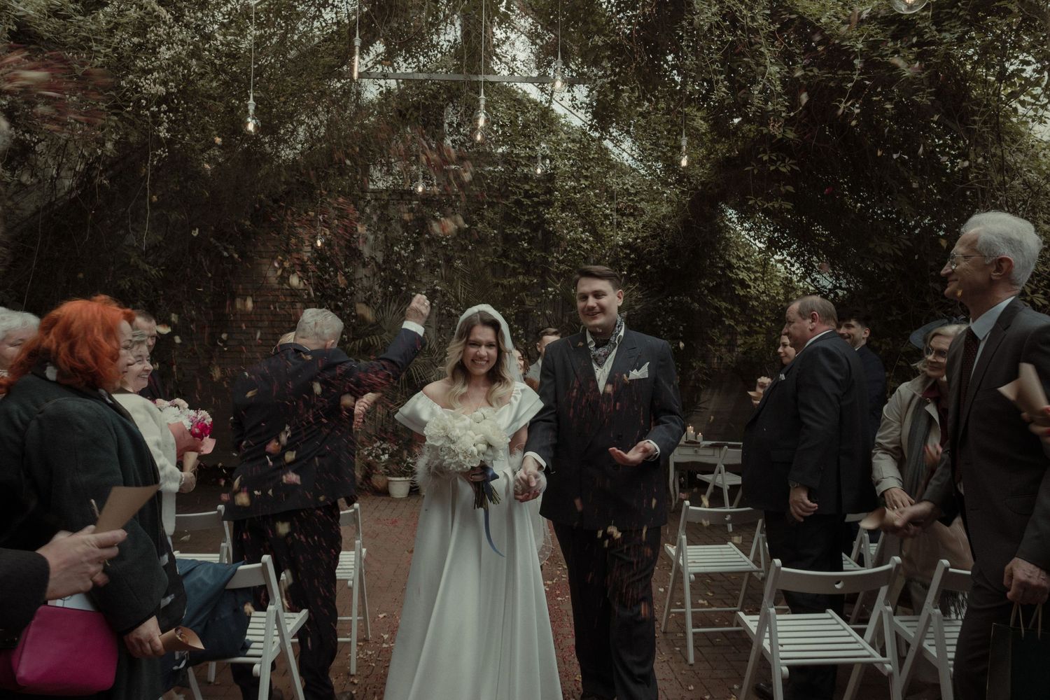 Wedding ceremony takes place outdoors under lush trees during golden hour with guests seated in white chairs.