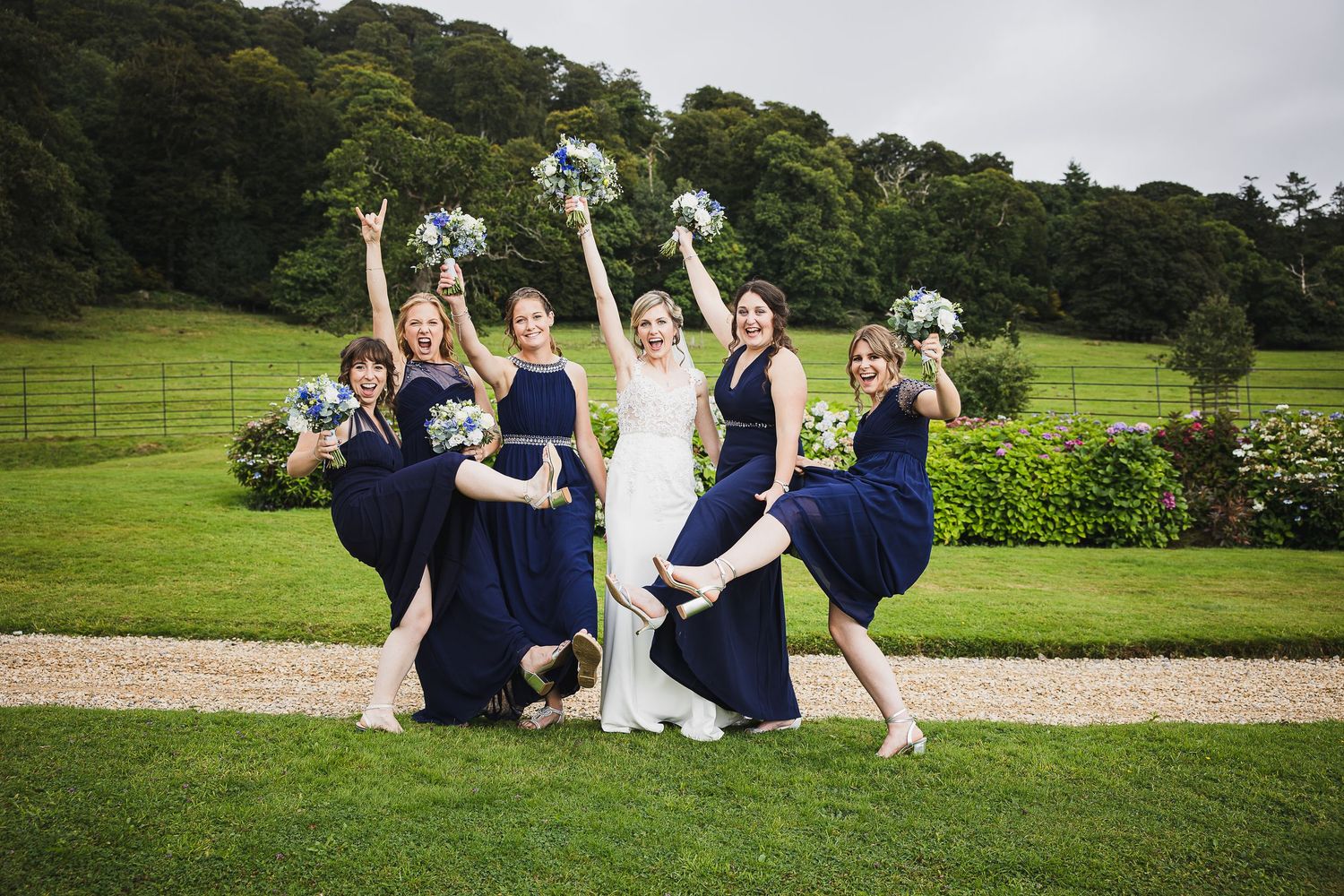 Wedding party in navy blue and white outfits pose playfully on a lush green lawn at a garden venue.