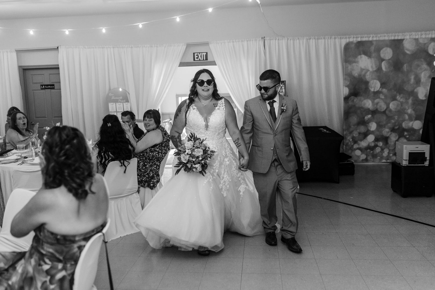 A black and white photo of a wedding ceremony with a couple walking together in a reception hall with string lights overhead.