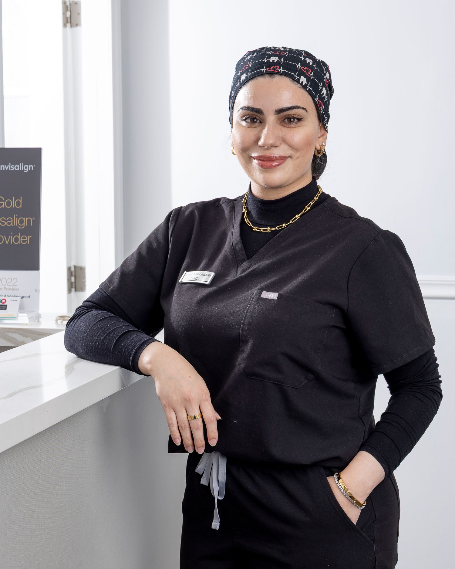 Healthcare worker in black uniform and patterned headwear poses in bright office setting.