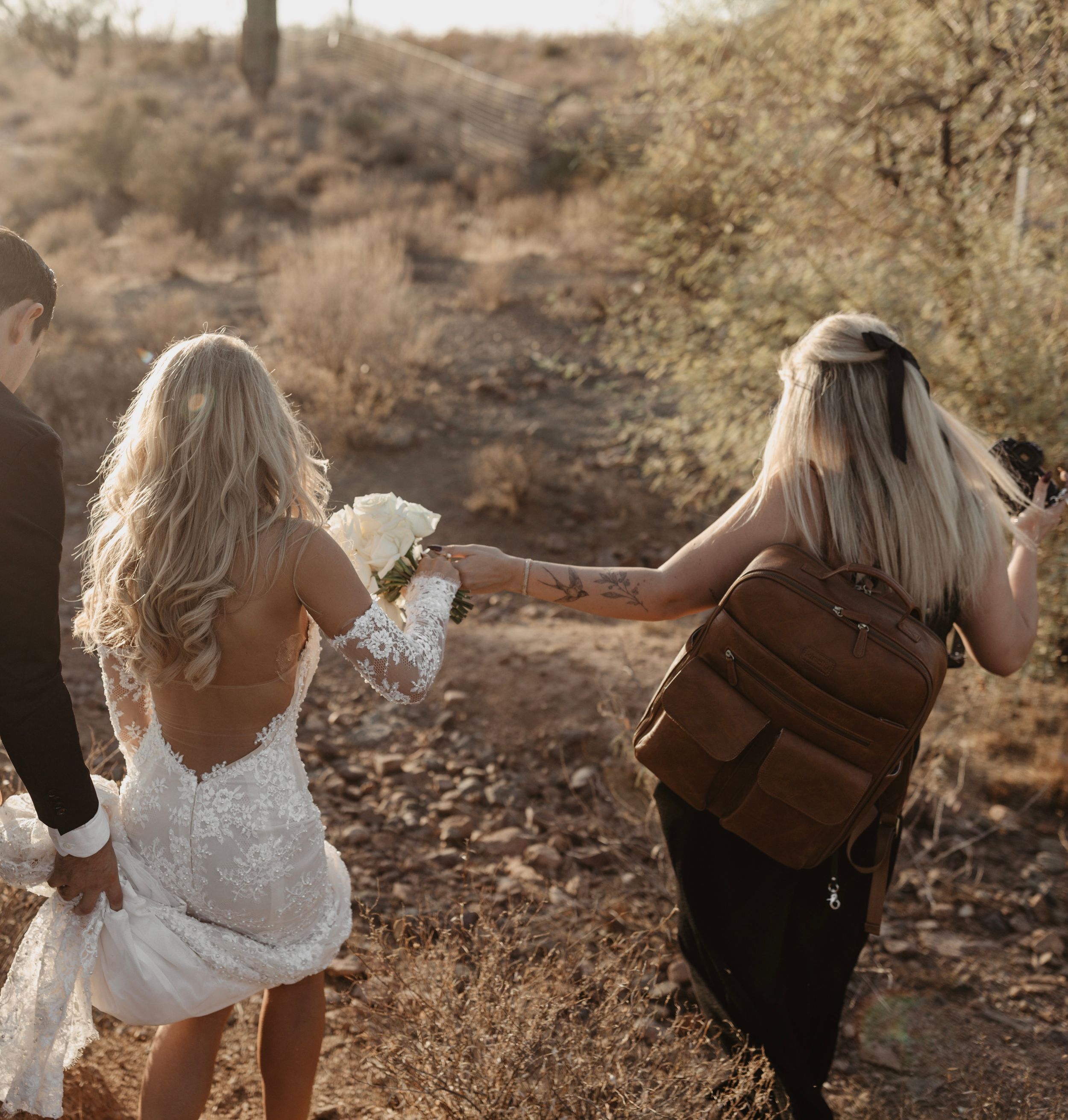 Wedding photographer with brown leather backpack follows couple walking along desert trail during golden hour sunset.
