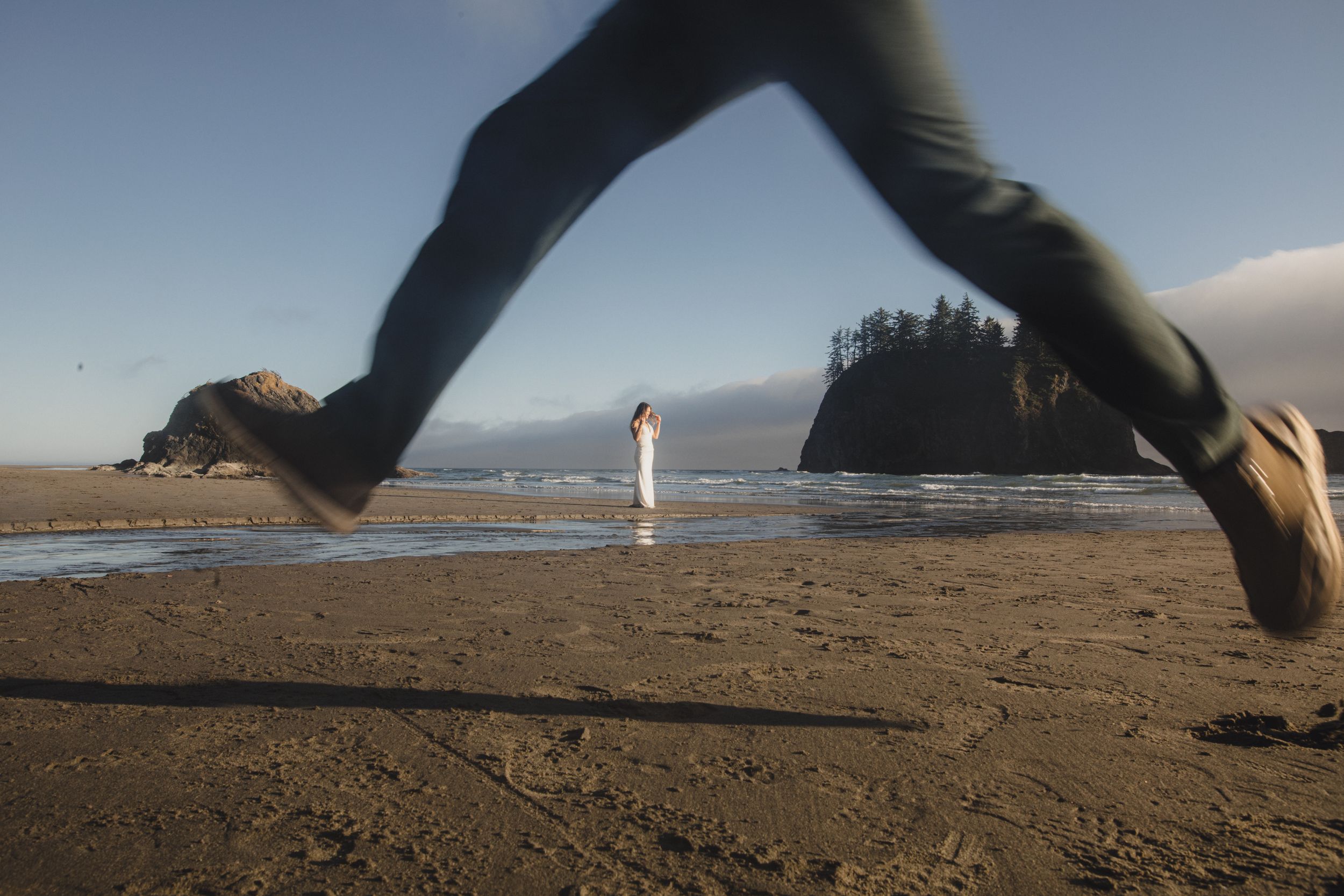 Motion blur of someone jumping on a beach with a distant sea stack and figure standing in the background.