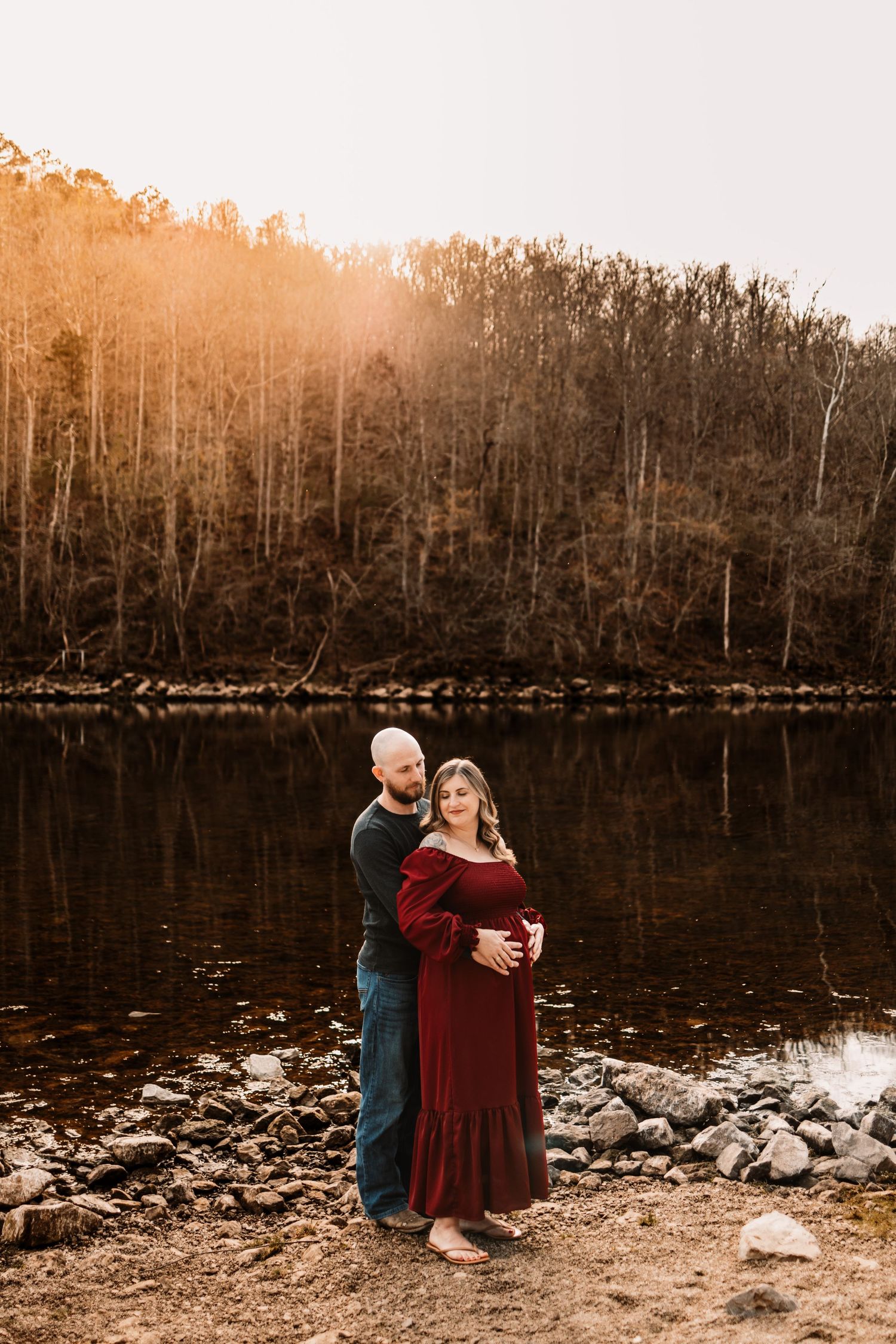 Couple embracing at golden hour by a reflective lake, pregnant woman in burgundy off-shoulder gown.