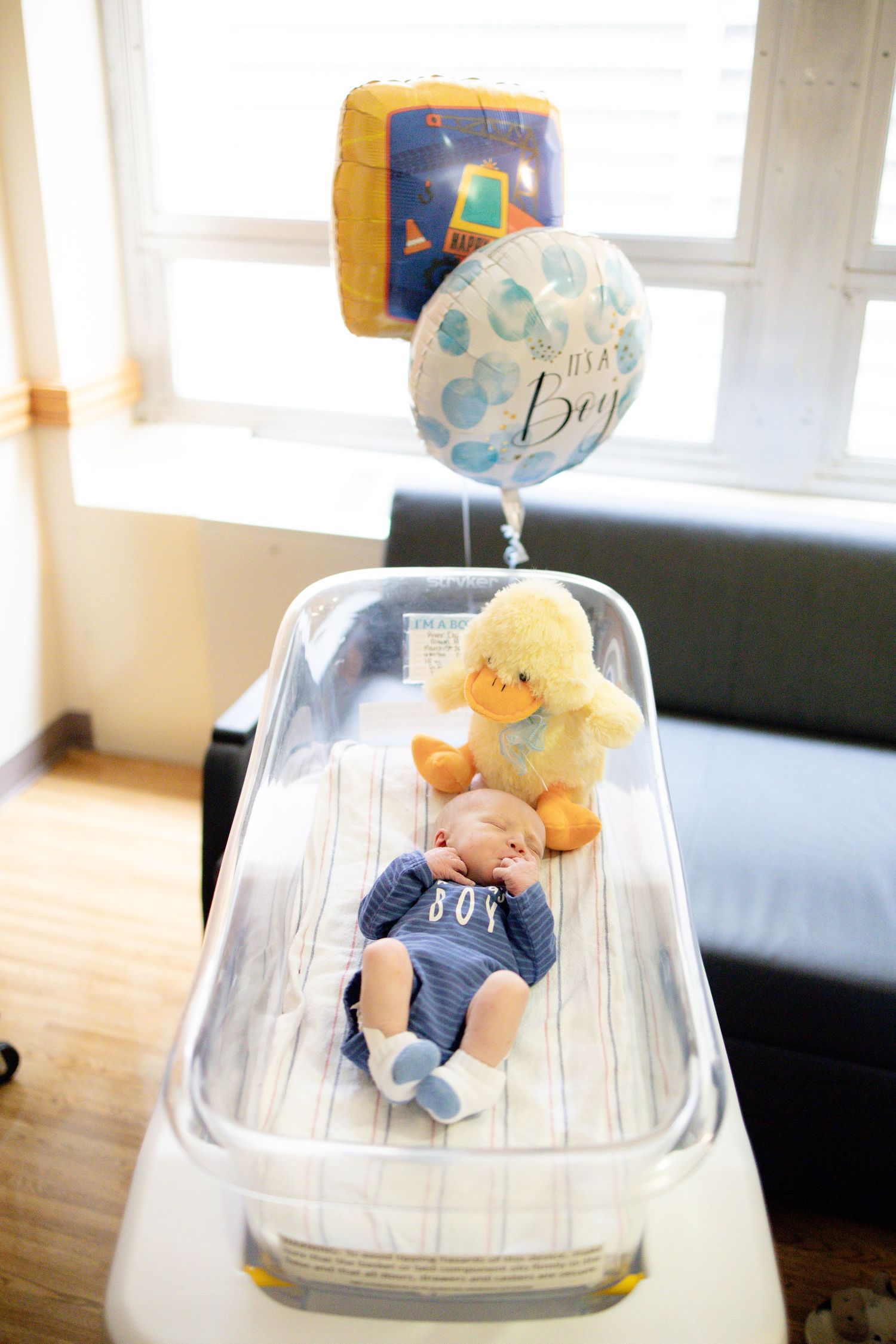 Newborn baby in blue outfit resting in hospital bassinet with It's a Boy balloon and duck plush toy.