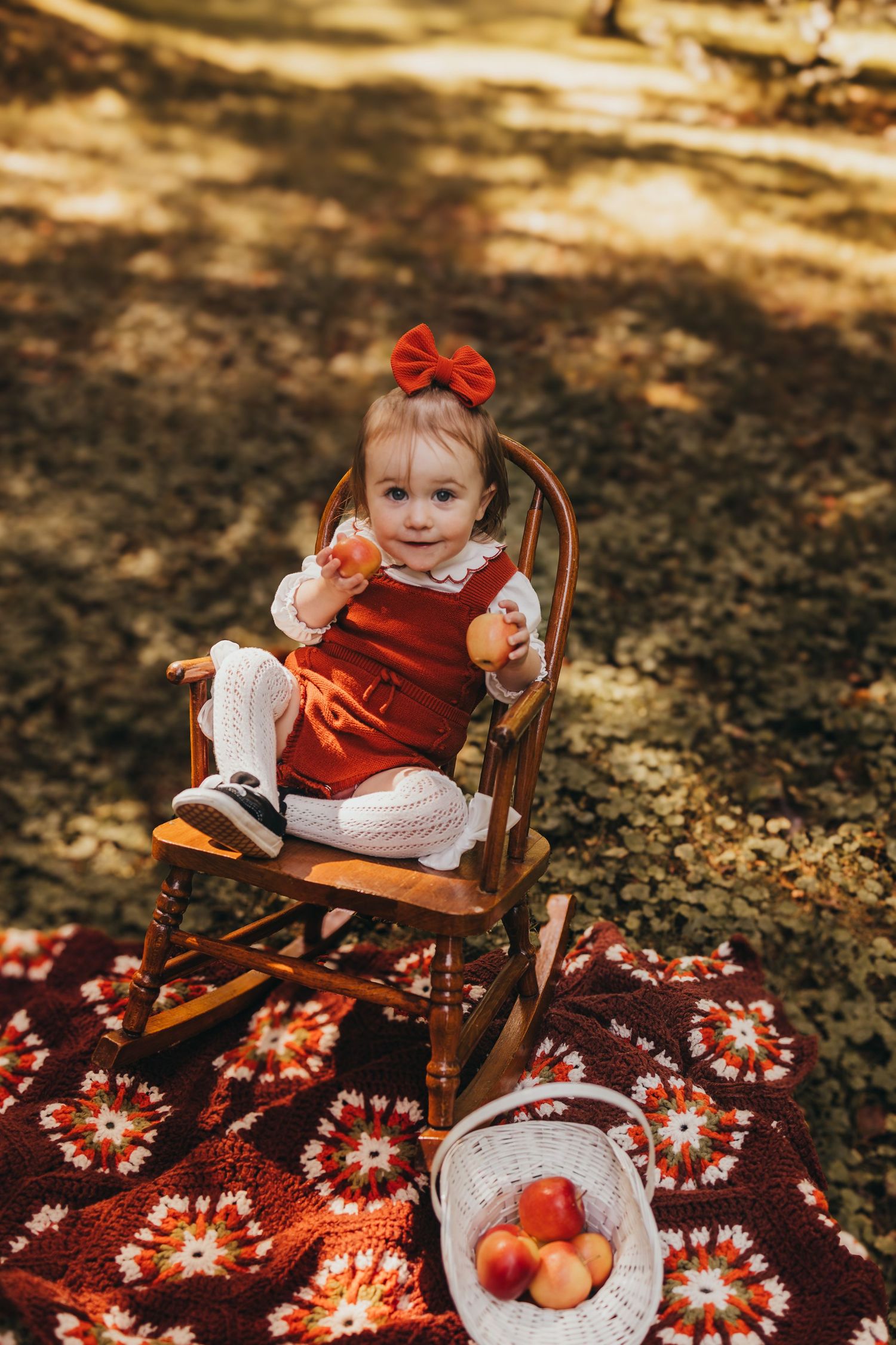 Toddler girl in red dress and bow sits in wooden rocking chair holding apple on colorful blanket outdoors.