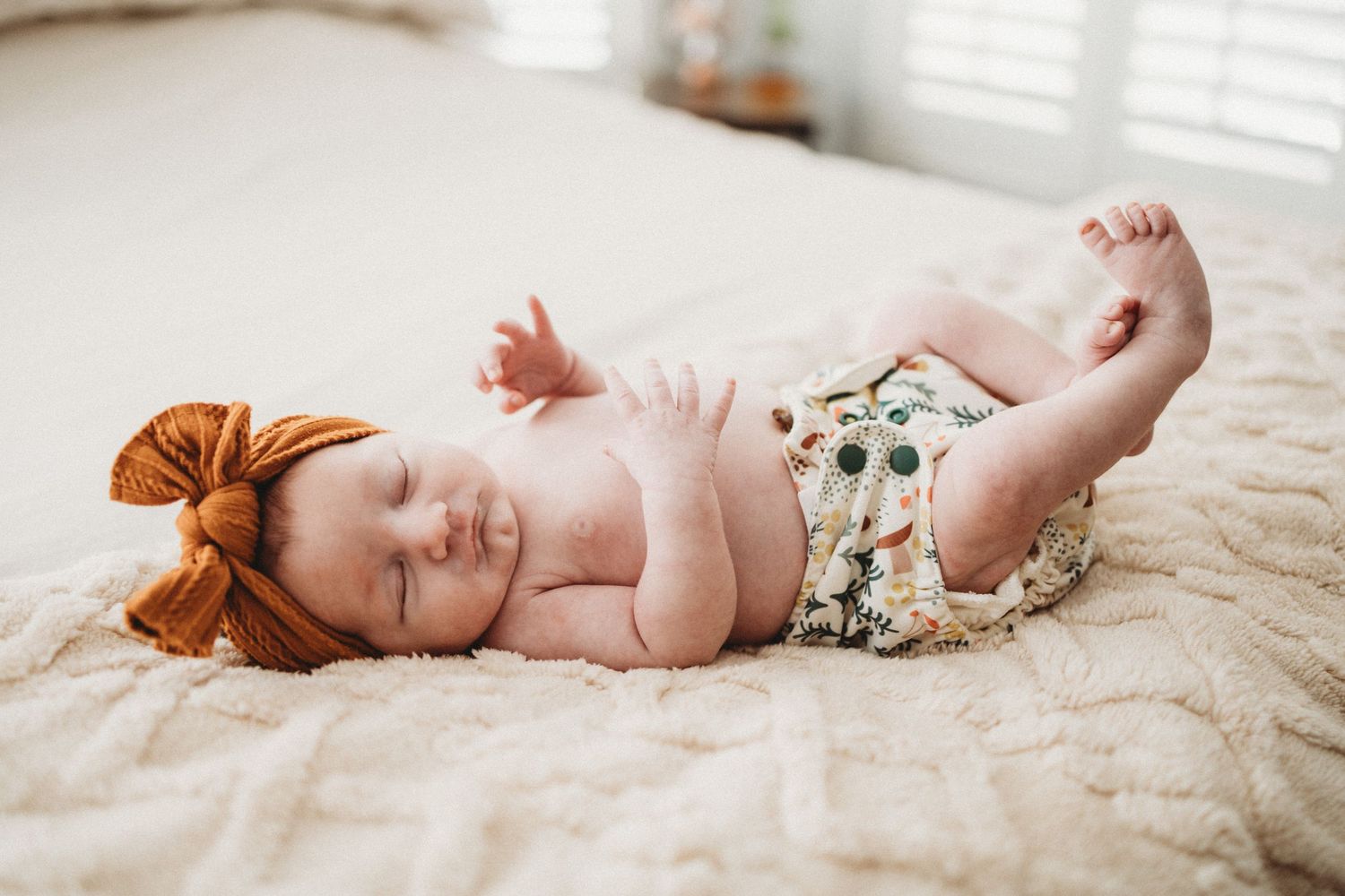 Peaceful newborn baby sleeping on cream blanket wearing floral diaper cover and rust bow headband.