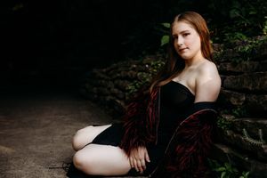 A figure in a flowing red dress poses gracefully among lush green foliage in an artistic outdoor photoshoot setting.
